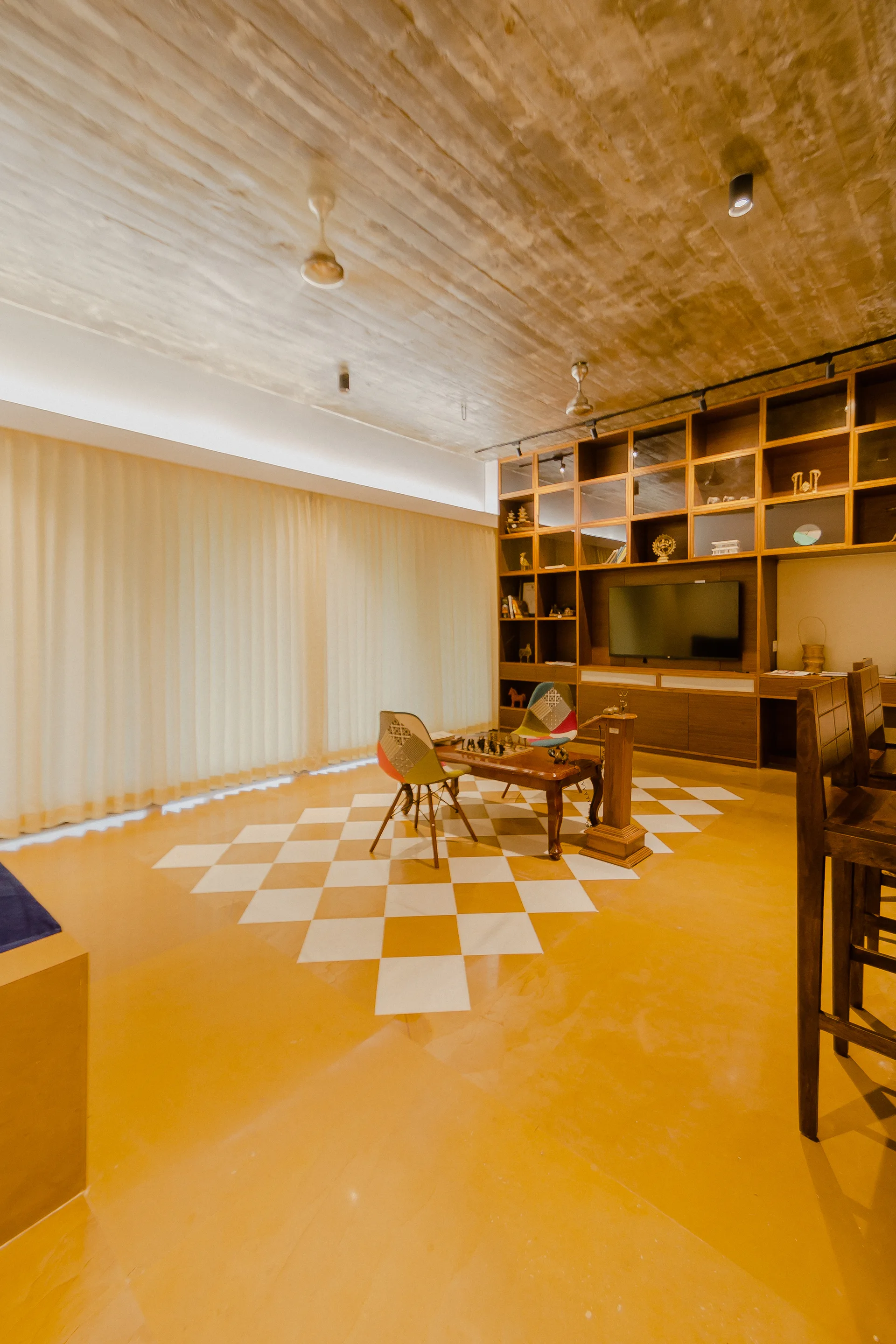 A living room features a yellow floor with a checkered tile inset, a large wooden built-in shelving unit, and a textured concrete ceiling.