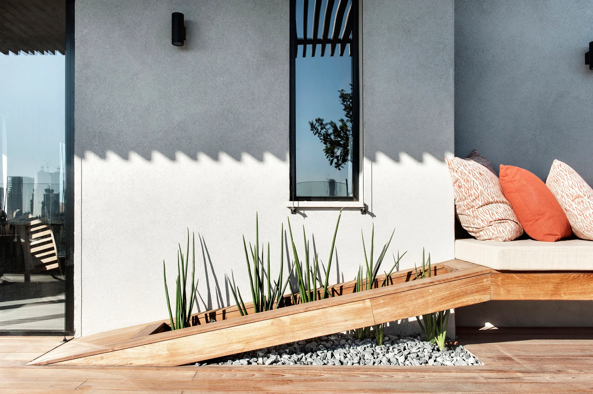 A modern wood patio deck features a built-in bench with orange throw pillows next to an integrated, angled wooden planter filled with gravel and green plants.
