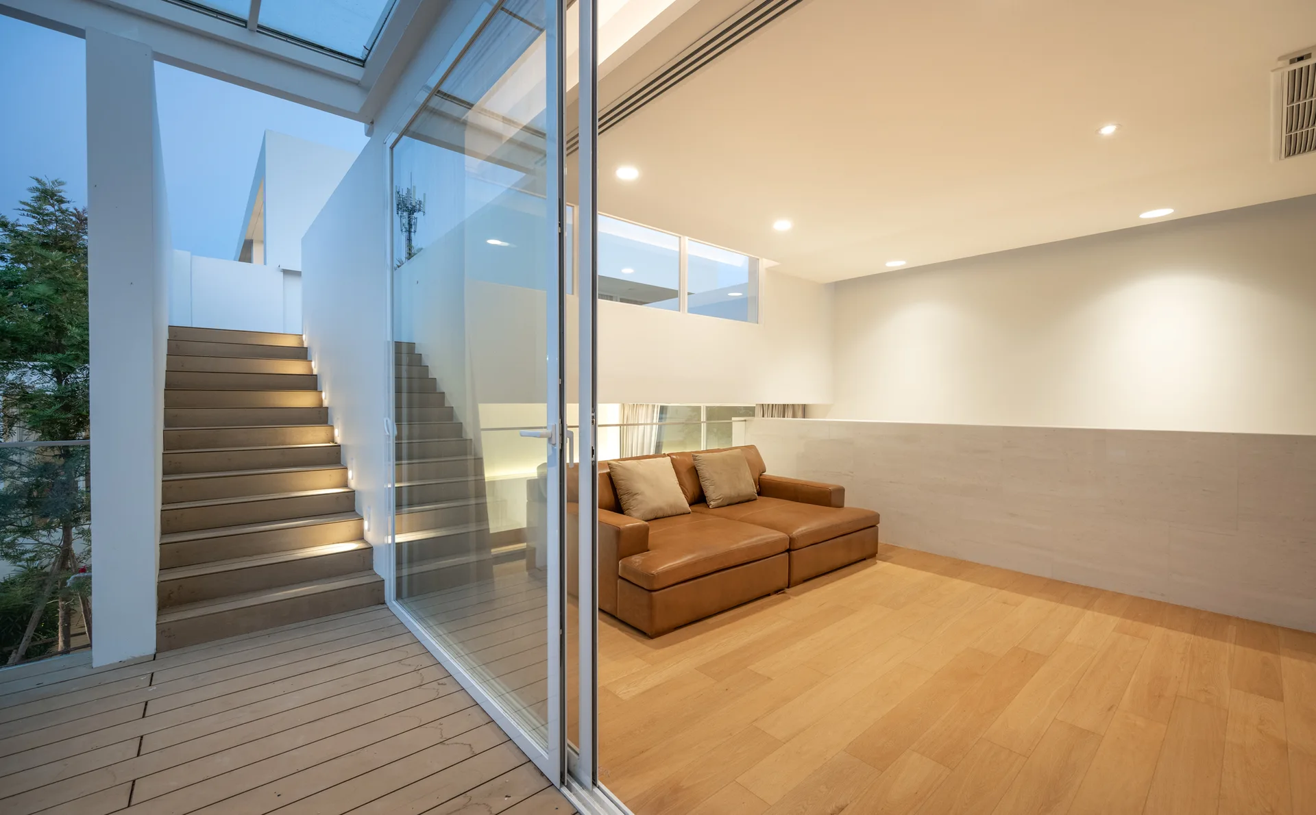 A modern living room features a brown leather sofa, light hardwood floors, and a sliding glass door opening to an illuminated outdoor staircase.