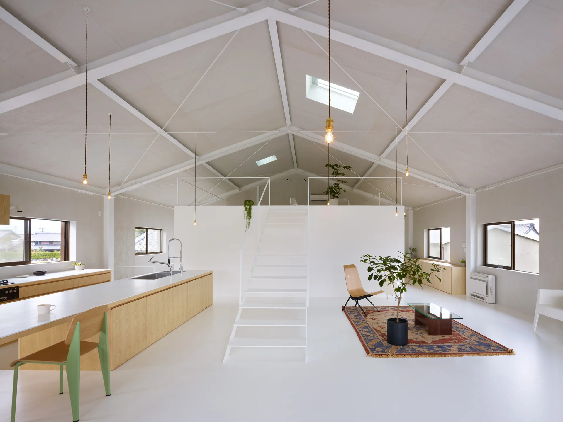 A bright minimalist open-plan loft featuring white vaulted ceilings, a central white staircase, a light wood kitchen island, and a patterned rug.