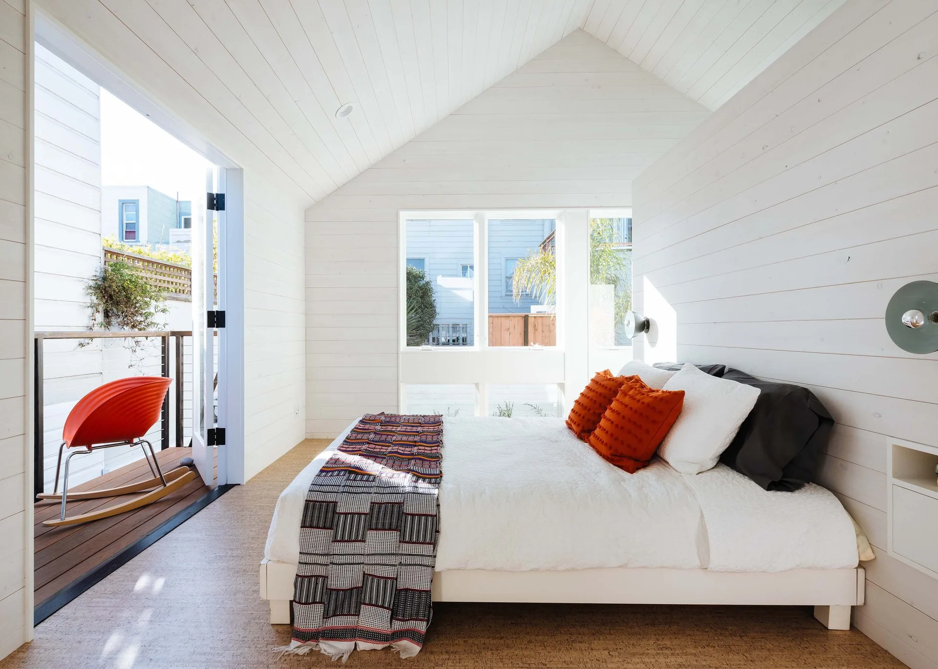 A modern white bedroom features a bed with orange pillows, shiplap walls, a vaulted ceiling, and an open door leading to a private wooden deck.