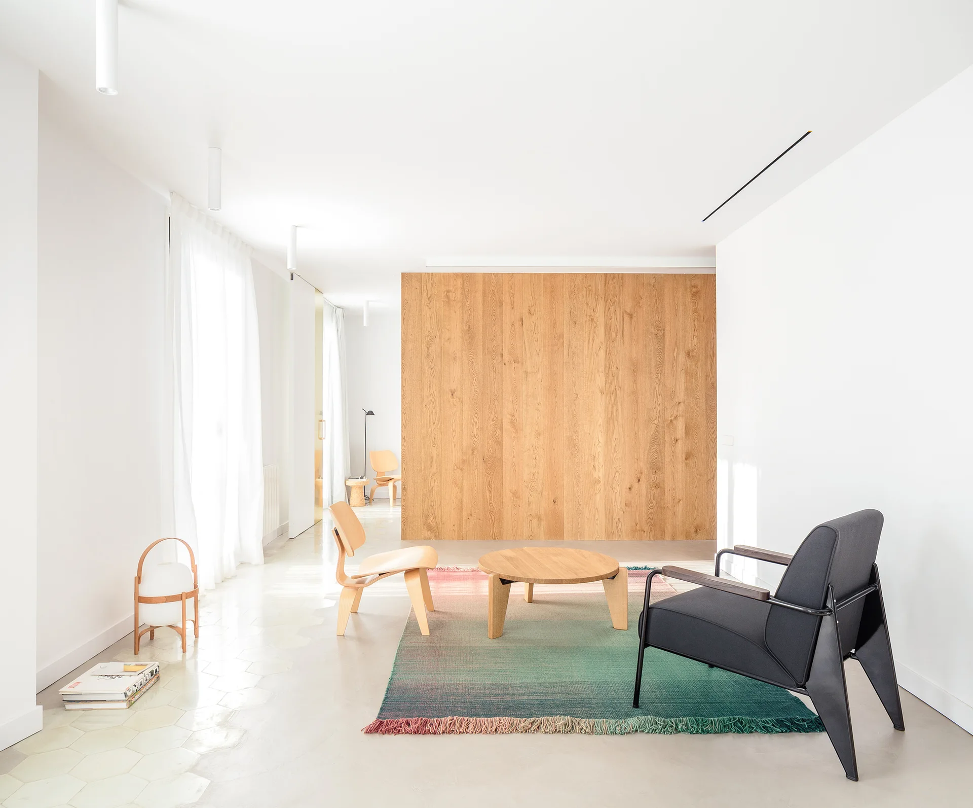A minimalist living room features a wooden accent wall, a circular coffee table on a colorful gradient rug, and a black armchair.