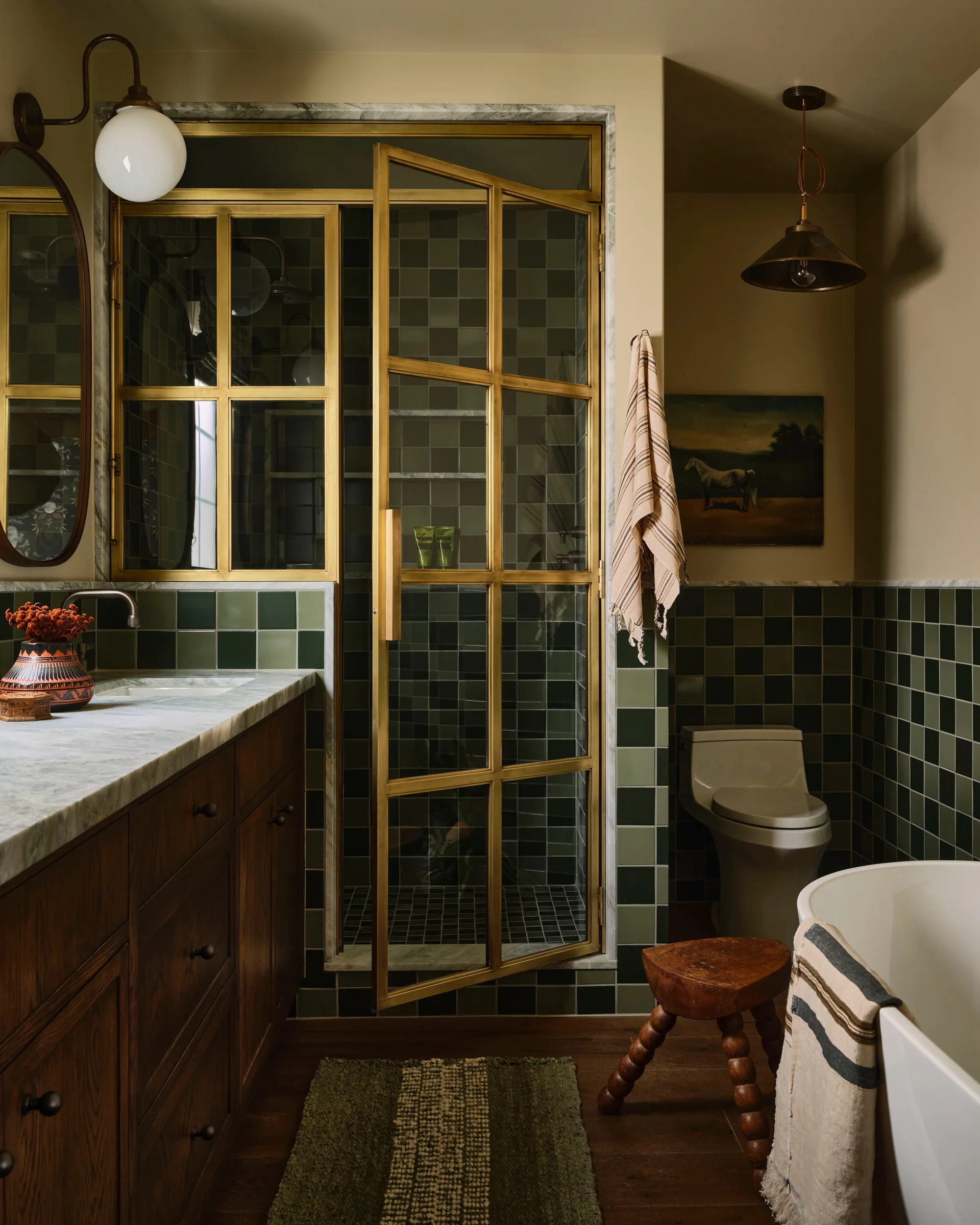 A bathroom featuring a dark wood vanity with a marble countertop, forest green gridded tile walls, and a brass-framed glass shower door.