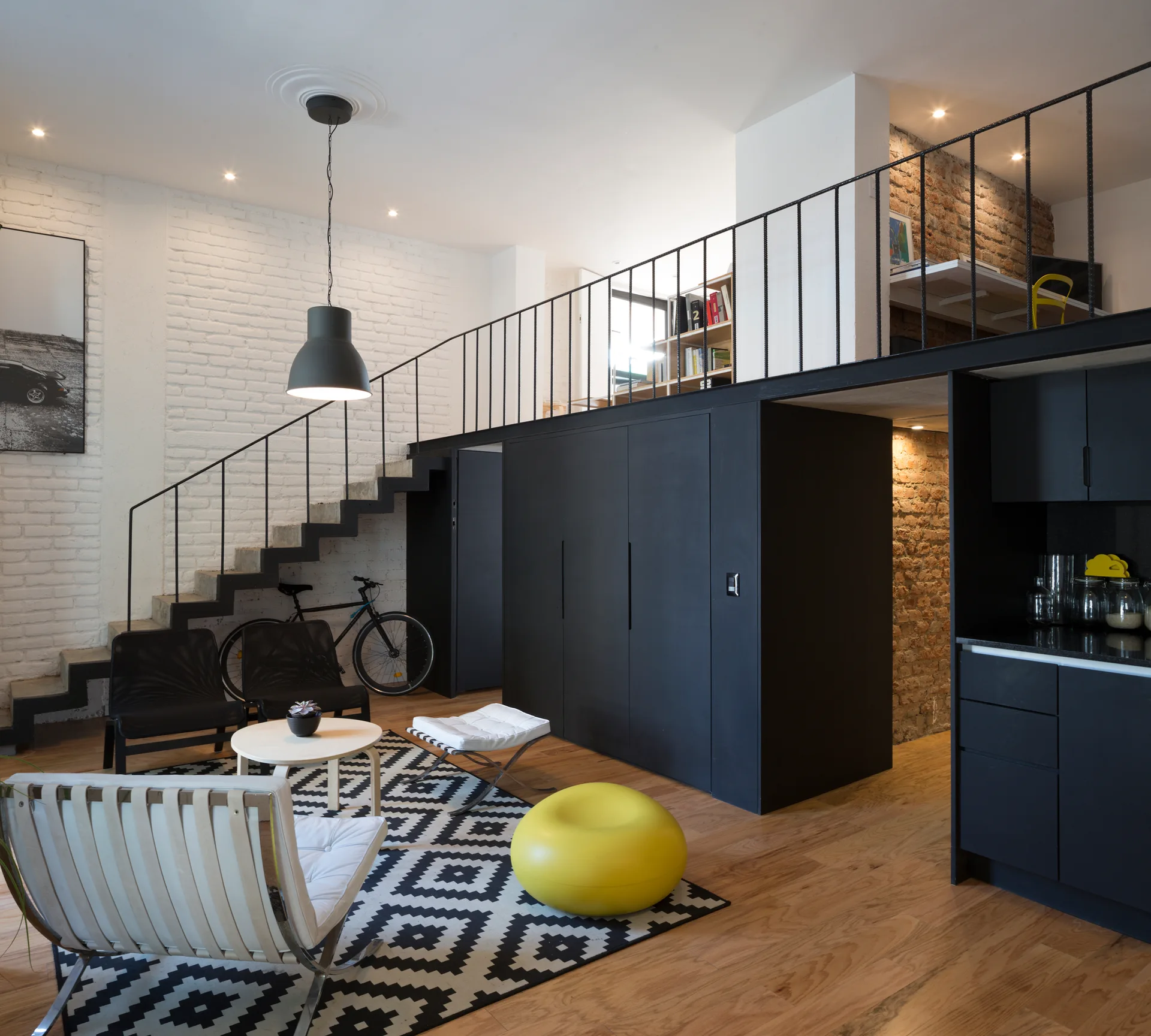 A modern loft living room featuring a black metal mezzanine, matching black under-loft storage, white brick walls, and a geometric pattern rug.