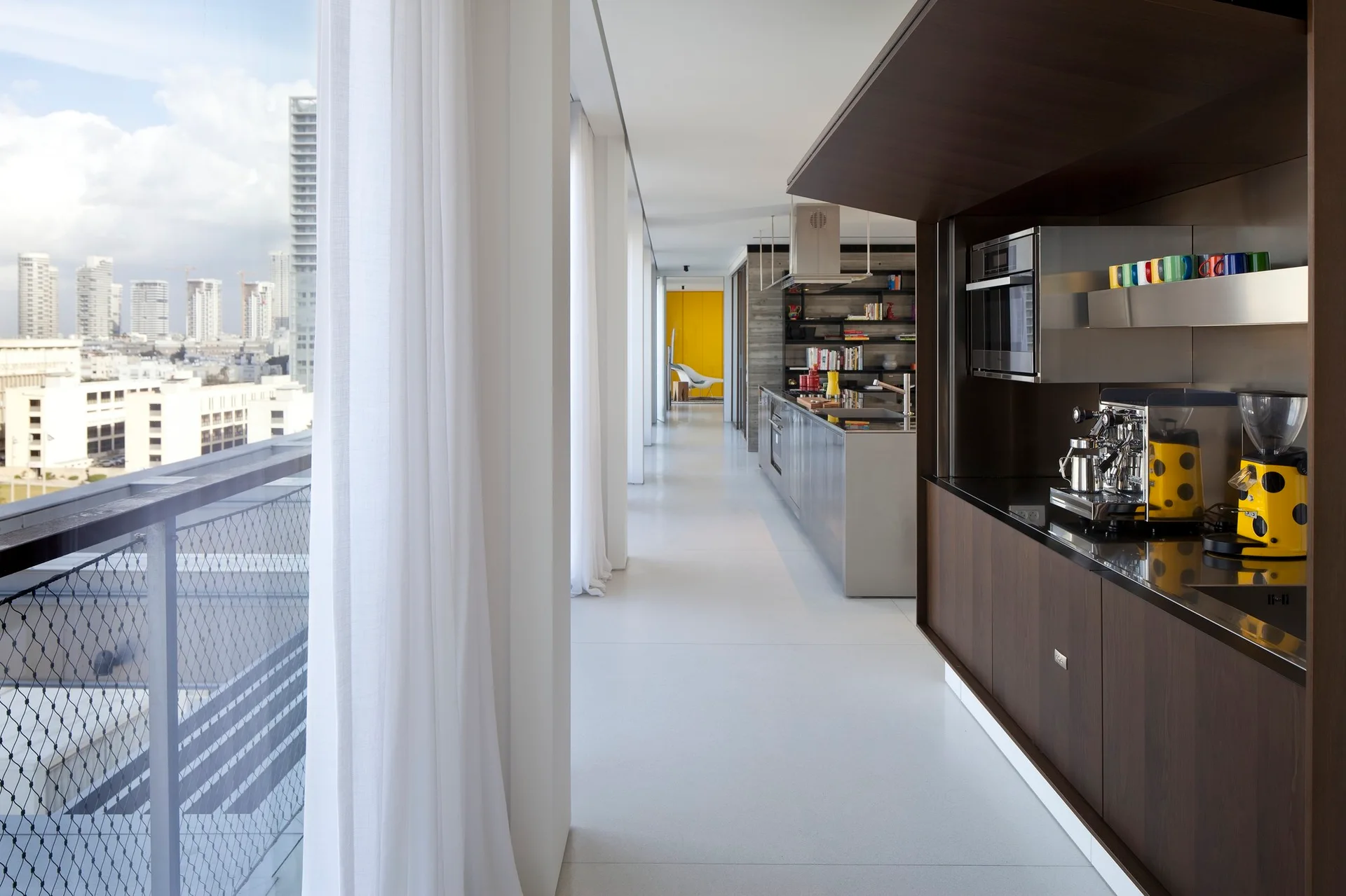 A long minimalist corridor kitchen with dark wood cabinets, a stainless steel island, and a bright yellow espresso machine next to floor-to-ceiling windows.