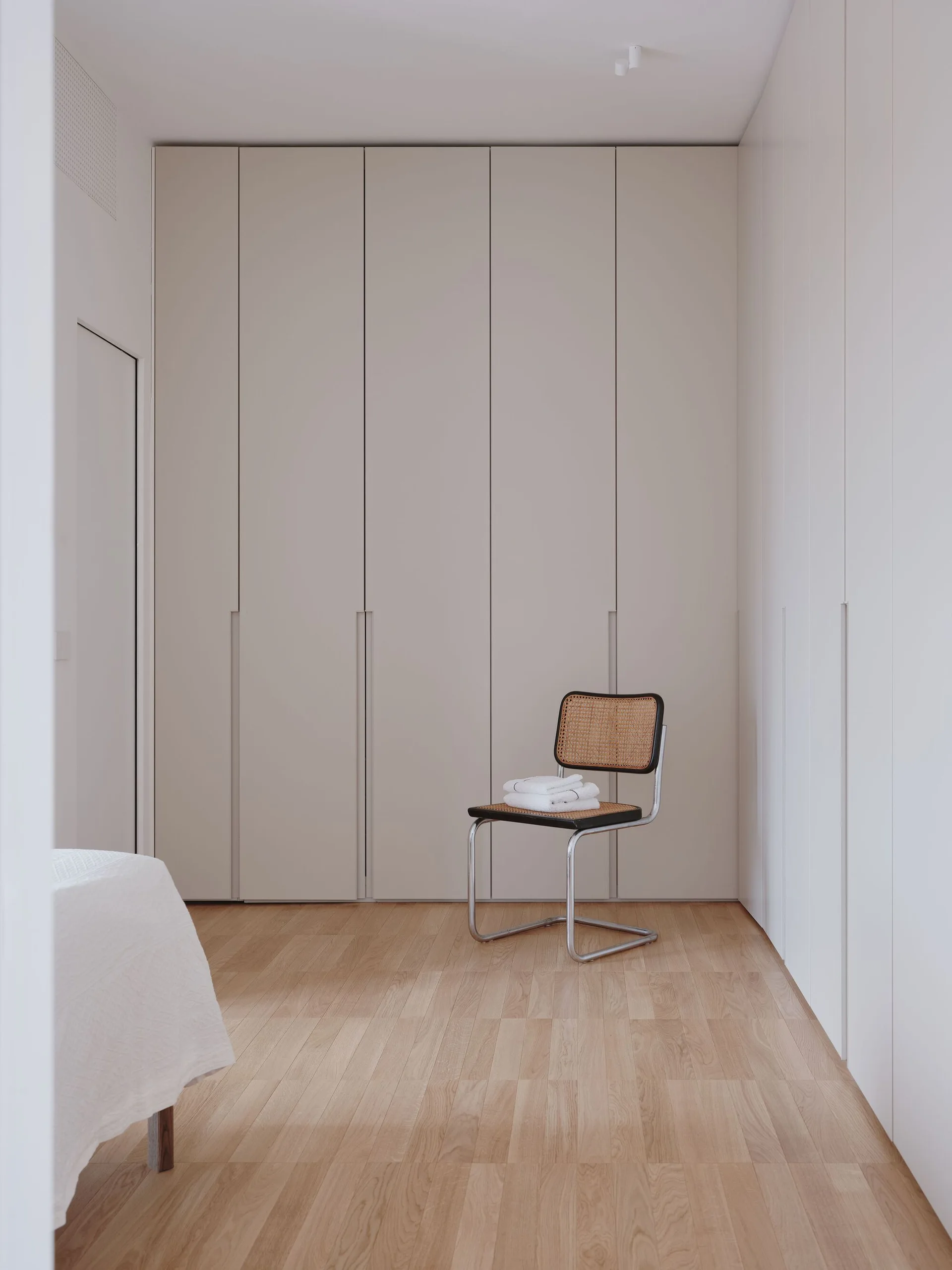 A minimalist dressing area with beige floor-to-ceiling built-in closets, light wood floors, and a single cane-backed chair holding folded white towels.