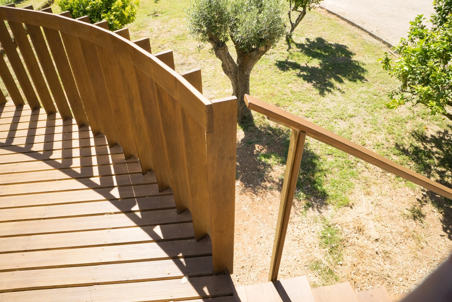 A curved wooden deck with a vertical slat railing and outdoor steps overlooks a sunlit grassy garden with trees.