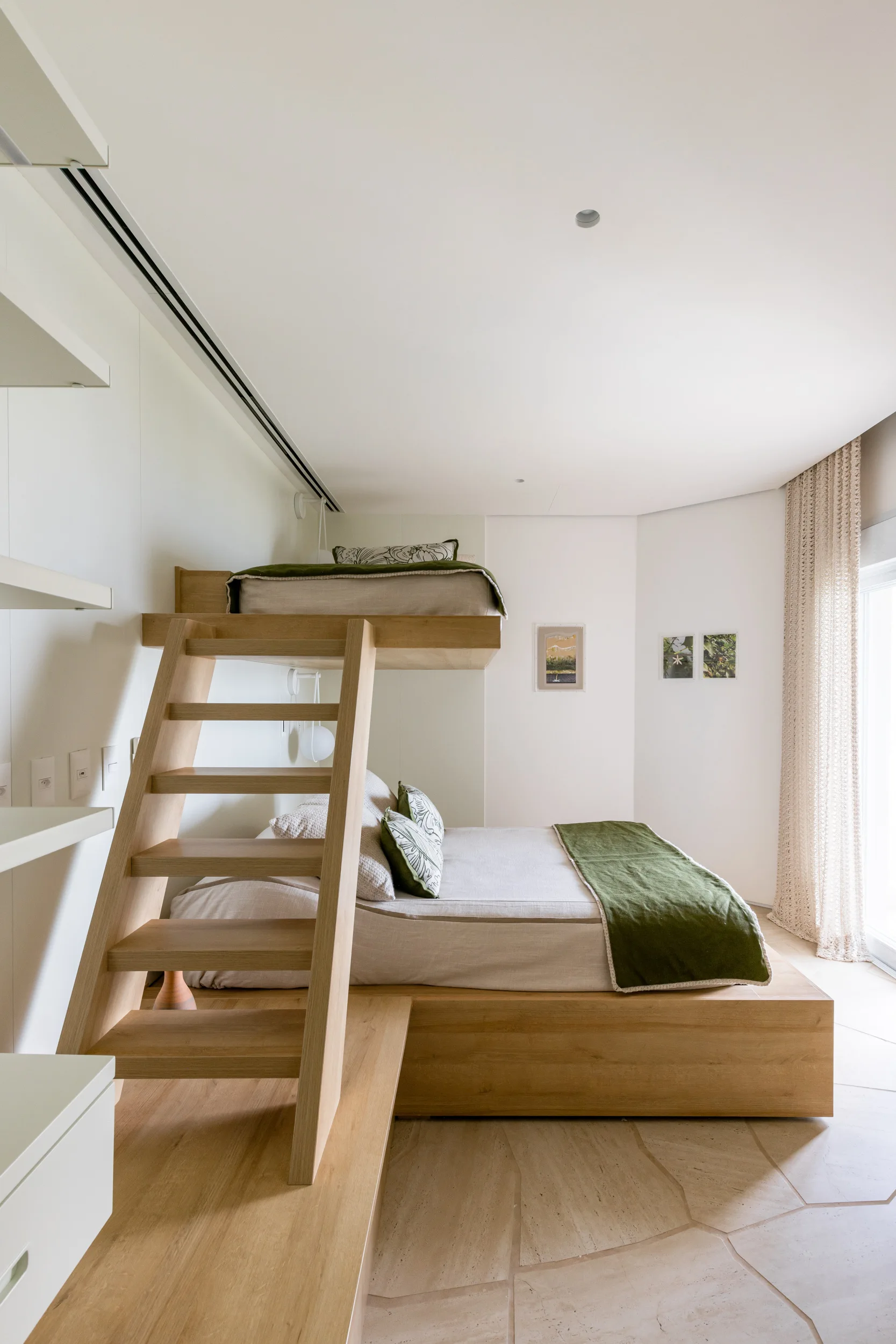 A modern bedroom features a custom wooden bunk bed structure with an integrated platform bed, olive green bedding, and white built-in wall shelving.