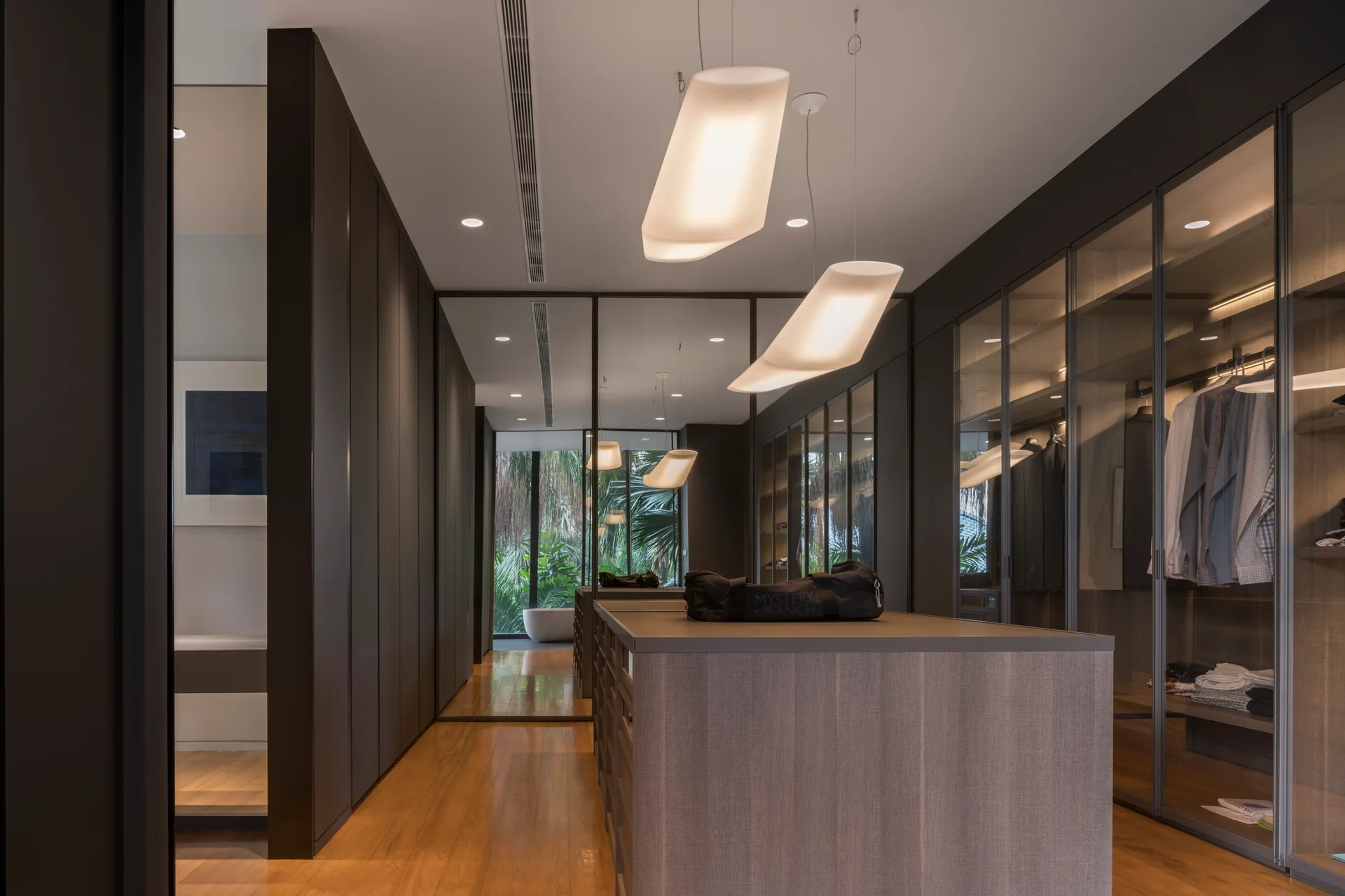 A modern walk-in closet with dark glass-front wardrobes, a central wood island, hardwood floors, and sculptural glowing pendant lights.