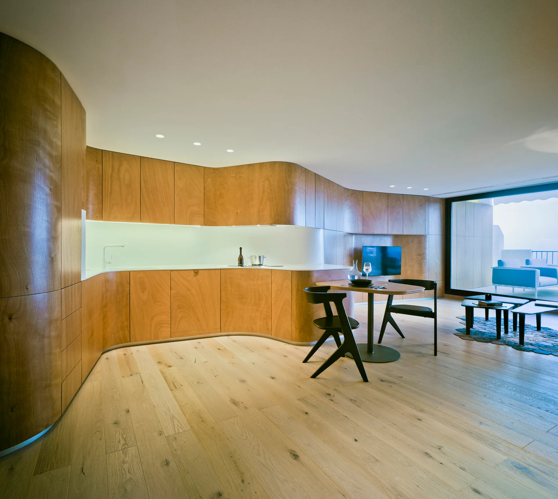 A minimalist open-plan kitchen features custom curved wood cabinetry, a round dining table with black chairs, and light wood flooring.