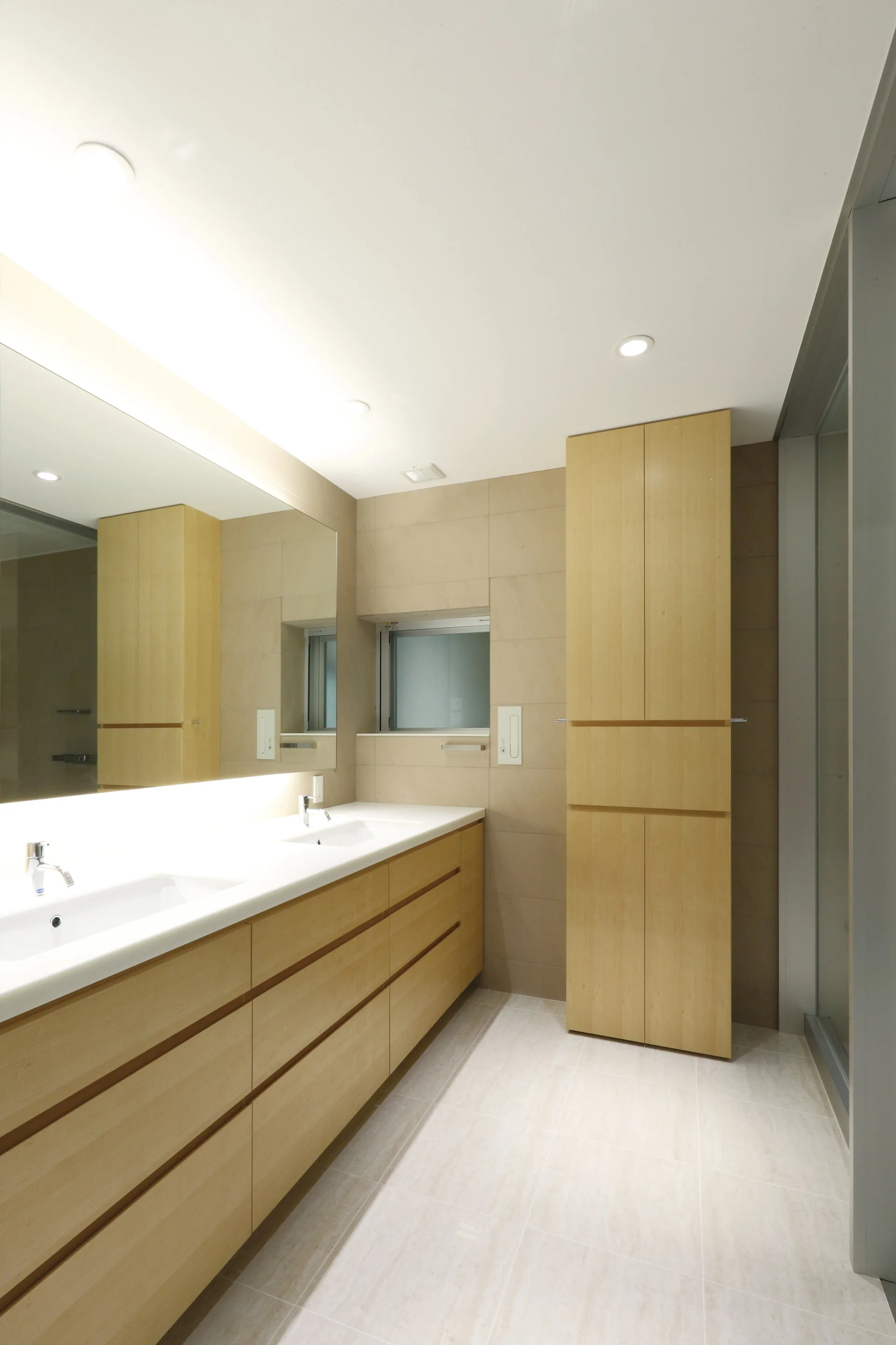 A modern minimalist bathroom featuring a long light wood double vanity, a matching tall linen cabinet, and a large backlit mirror against beige tile.