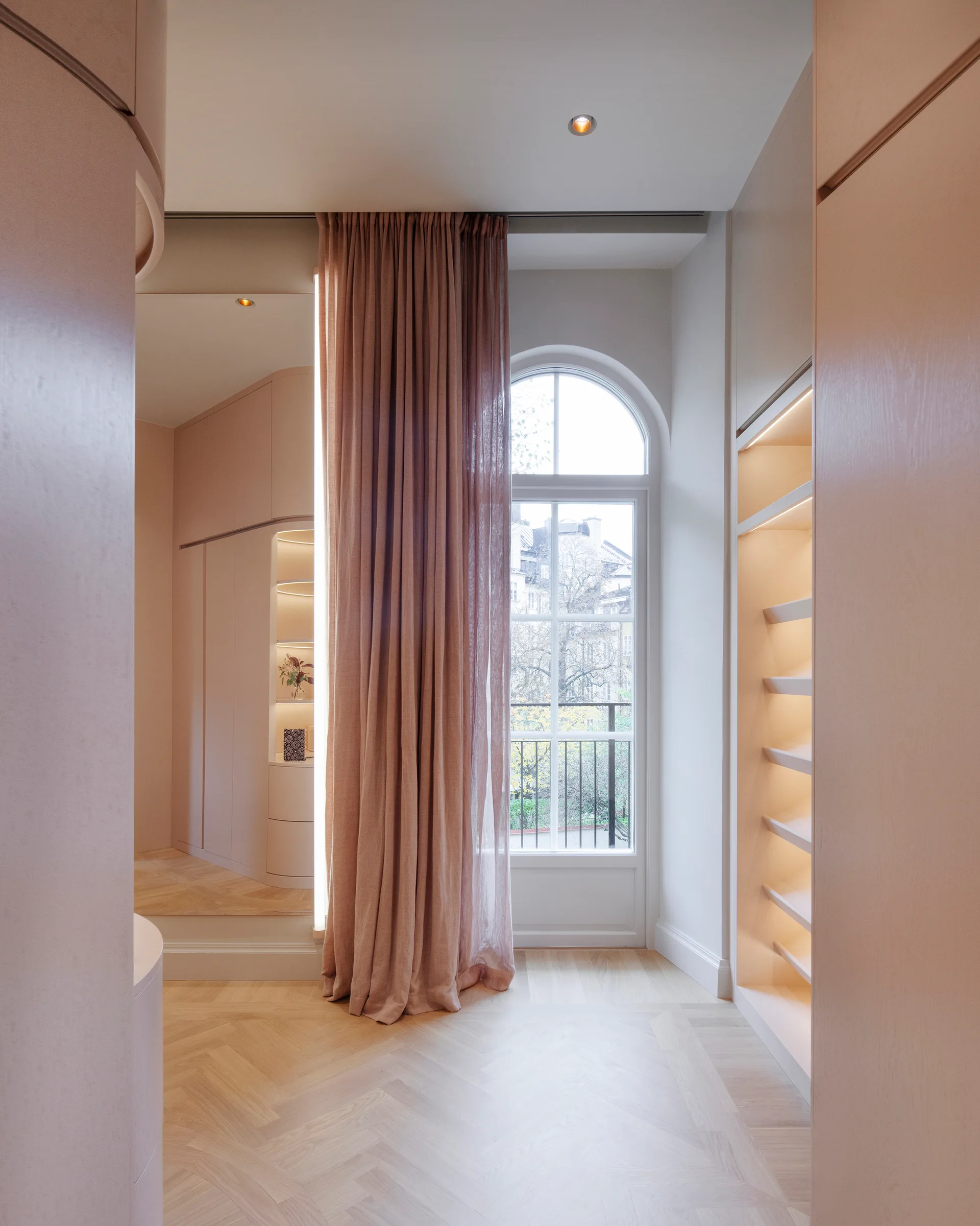A dressing room features custom light wood wardrobes, illuminated shelves, herringbone flooring, and an arched window draped with a long pink curtain.