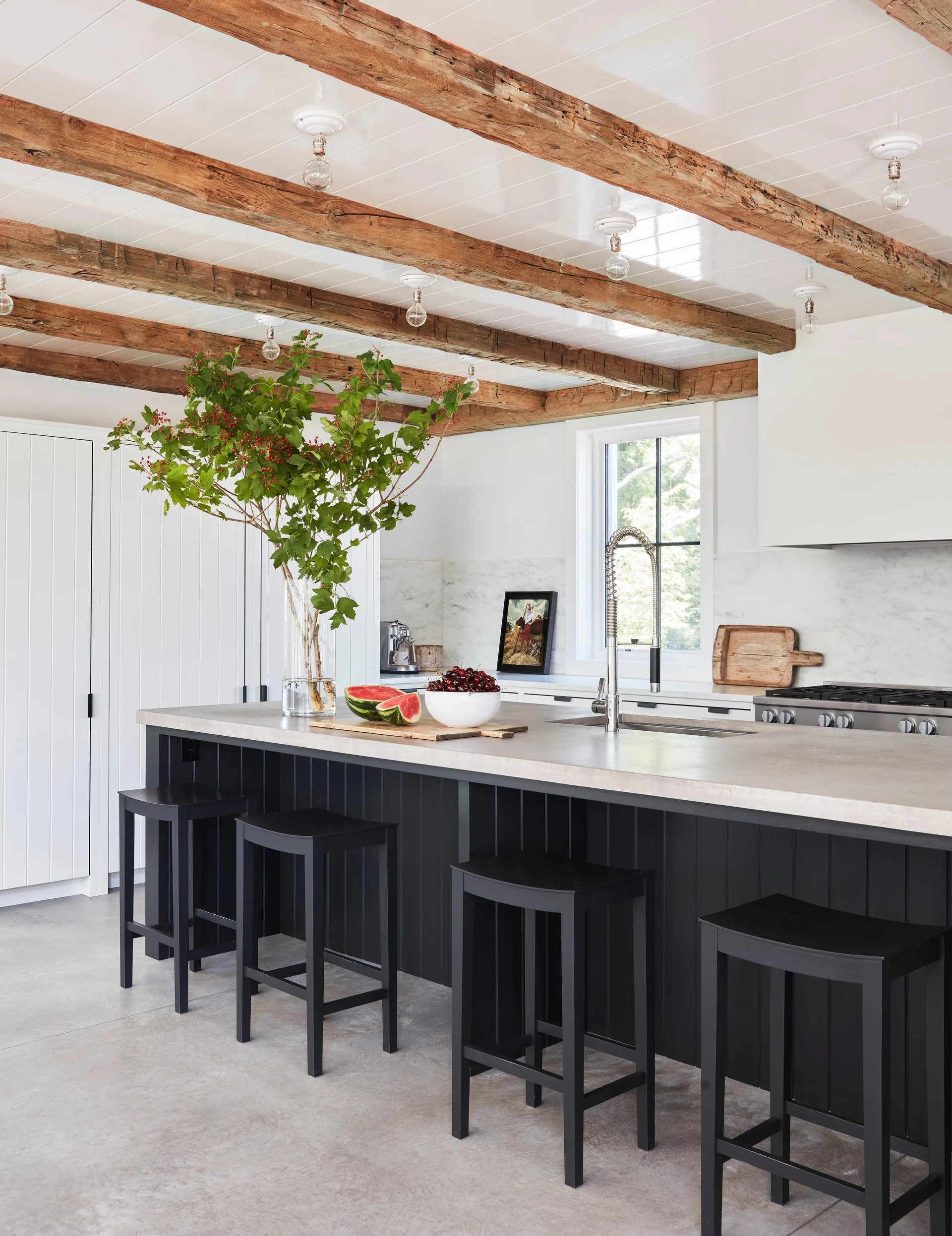 A modern farmhouse kitchen featuring a large black paneled island with bar stools beneath rustic exposed wood ceiling beams.