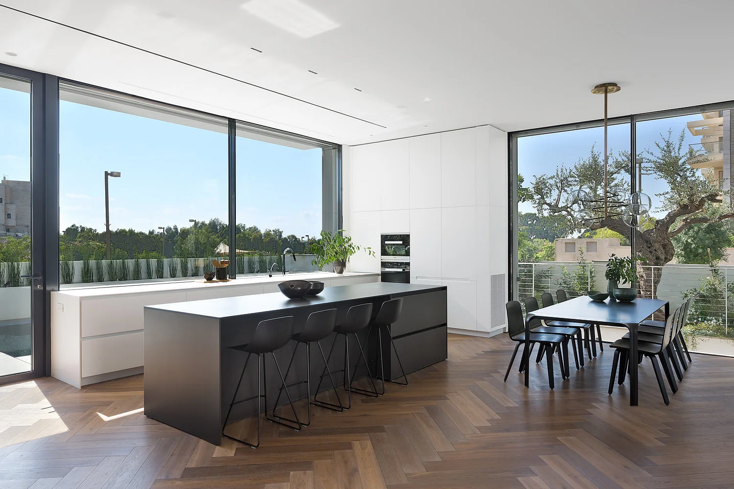 A modern open-plan kitchen features a large matte black island with stools, a matching dining table, and herringbone wood floors near large windows.