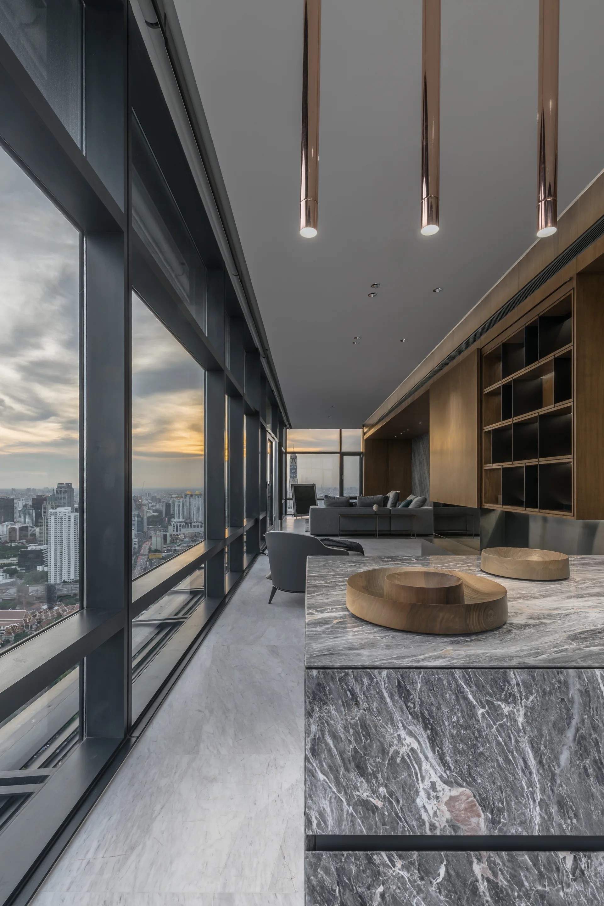 A modern interior features a grey marble kitchen island with wooden bowls, sleek copper pendant lights, and floor-to-ceiling windows showing a city skyline.