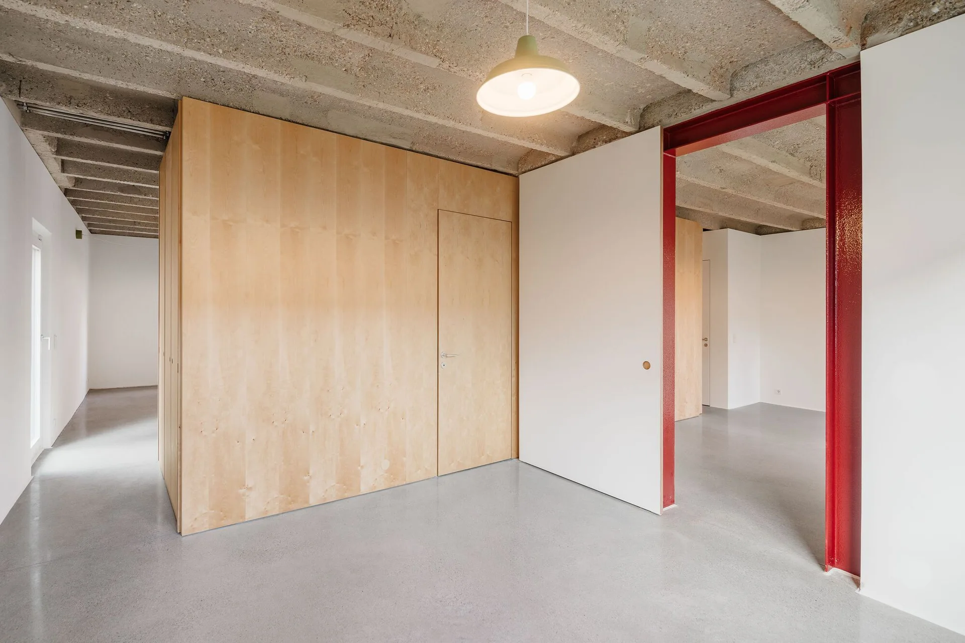A minimalist room features plywood wall paneling, a flush door, an exposed raw concrete ceiling, and a bright red structural steel beam.