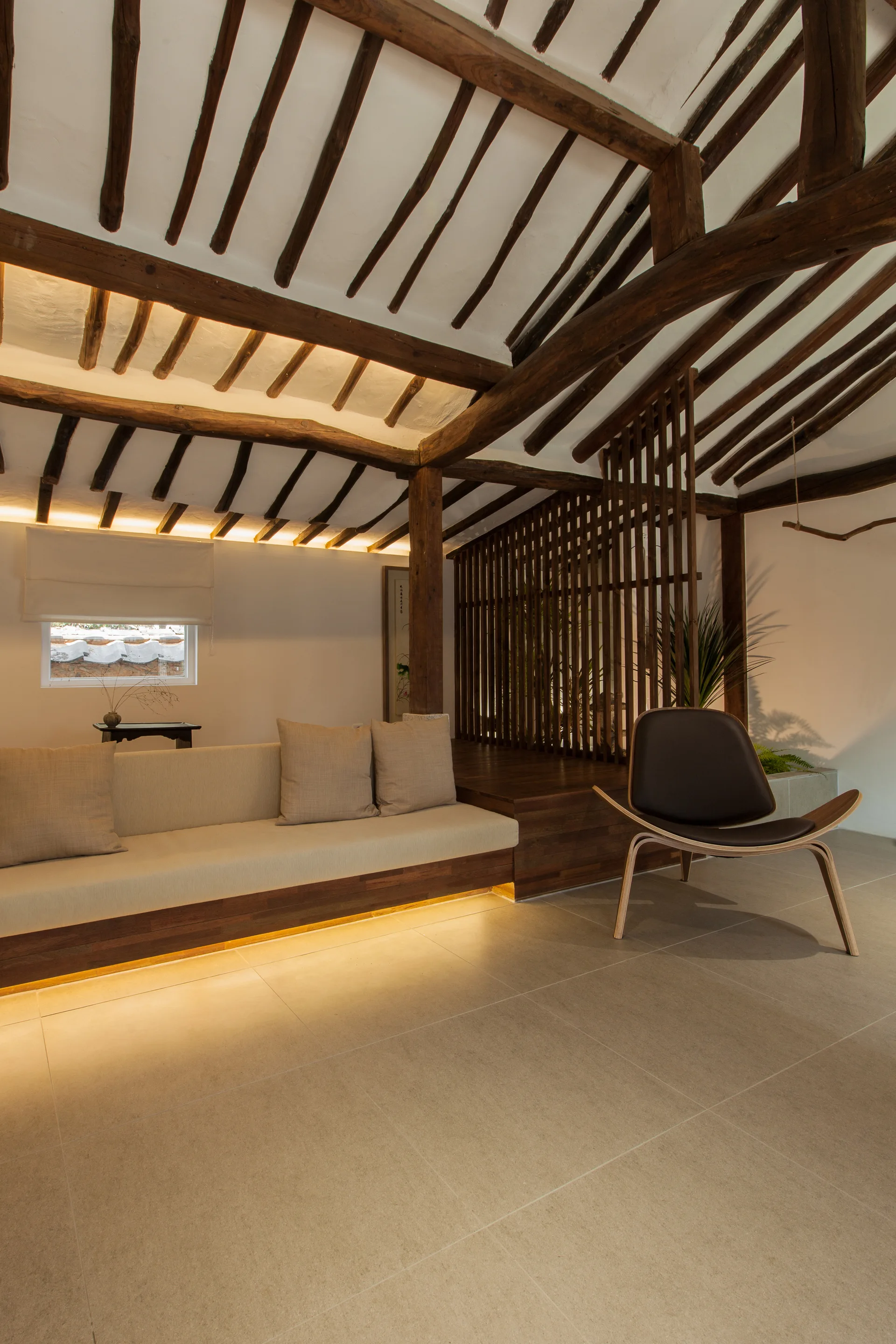 A minimalist room featuring exposed wood ceiling rafters, a low built-in sofa on a wood platform with under-bench lighting, and a curved lounge chair.
