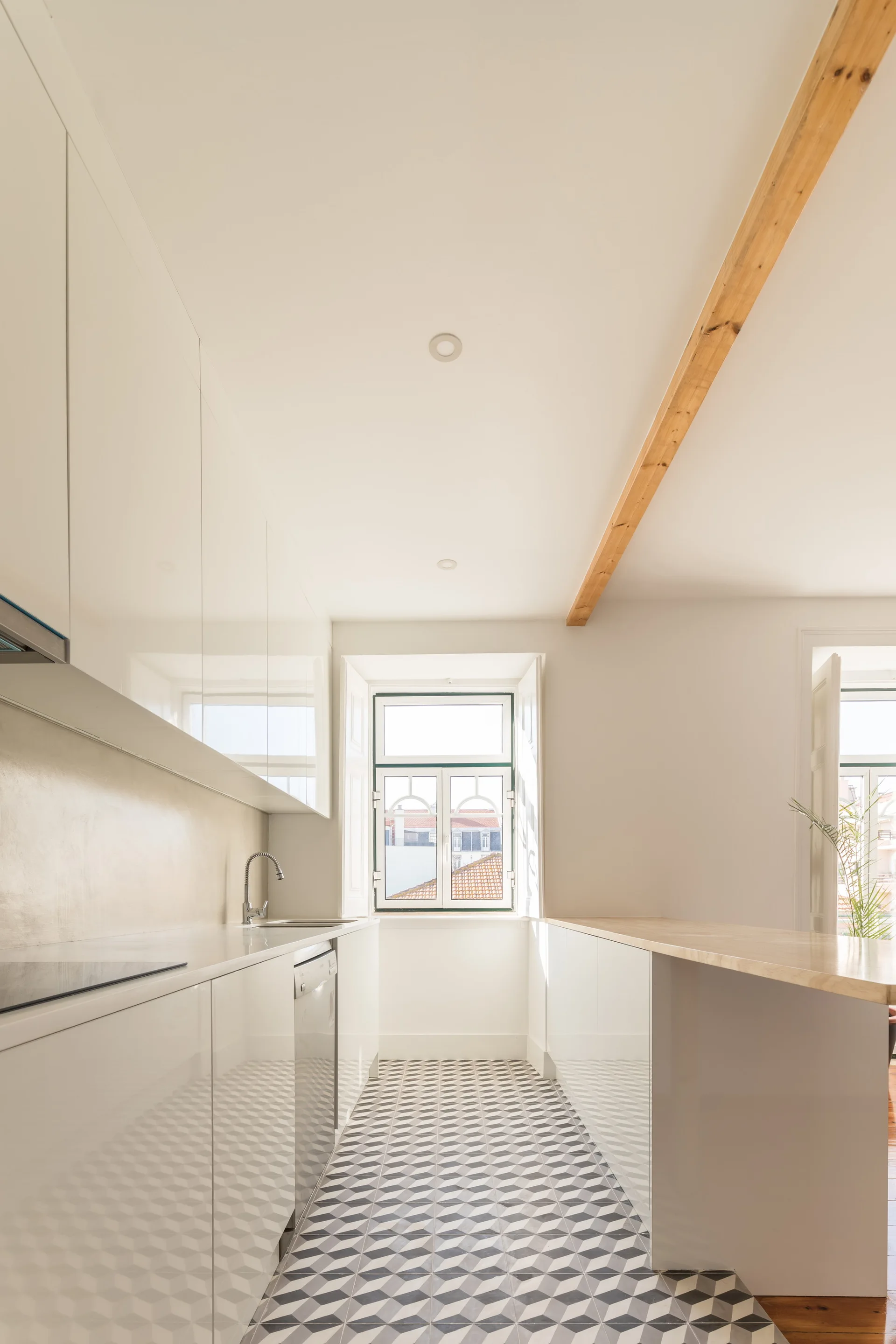 A narrow galley kitchen featuring glossy white flat-panel cabinets, geometric patterned floor tiles, and a natural wood ceiling beam.