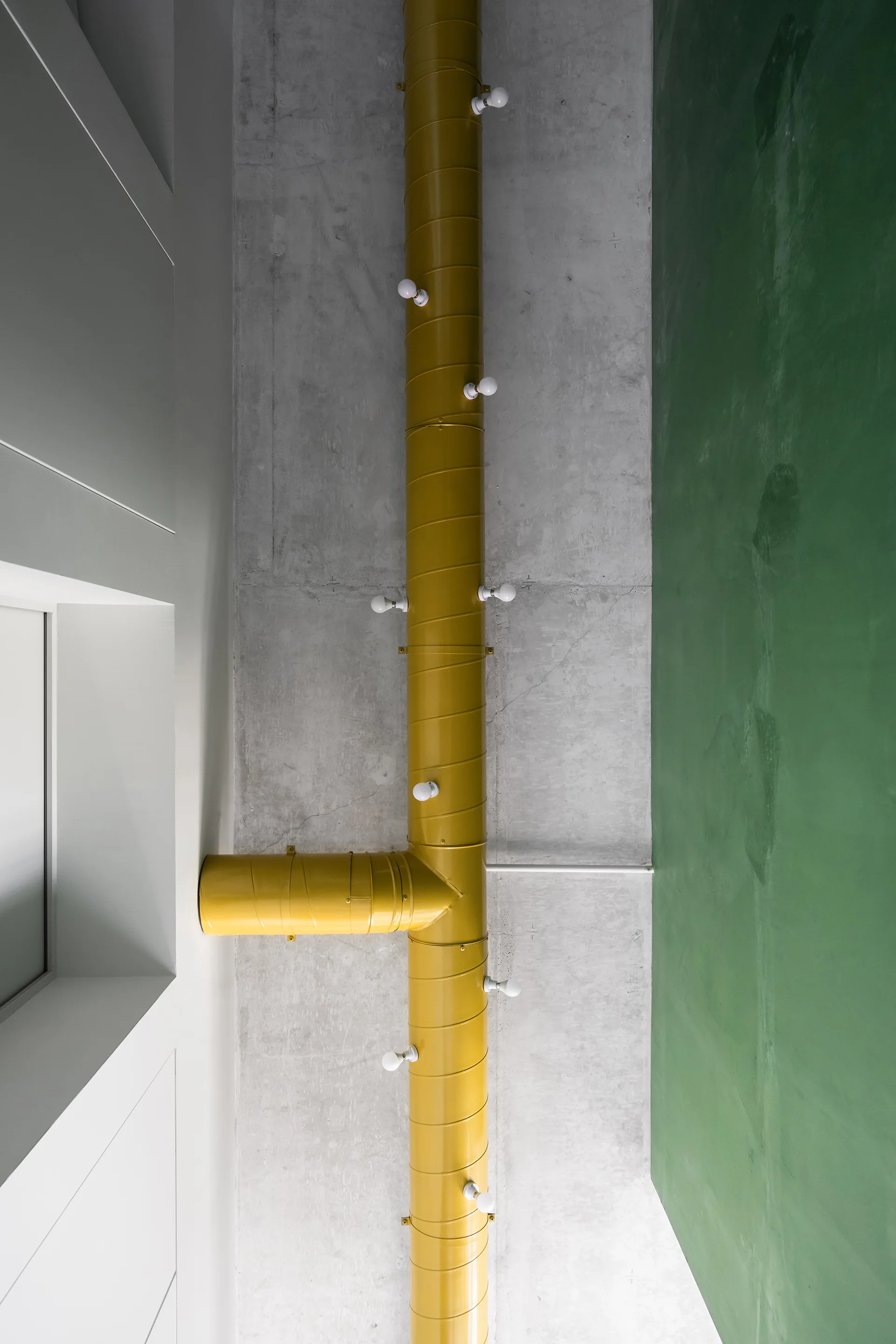 A T-shaped yellow ventilation duct with small light bulbs mounted on an exposed concrete ceiling next to a solid green painted surface.