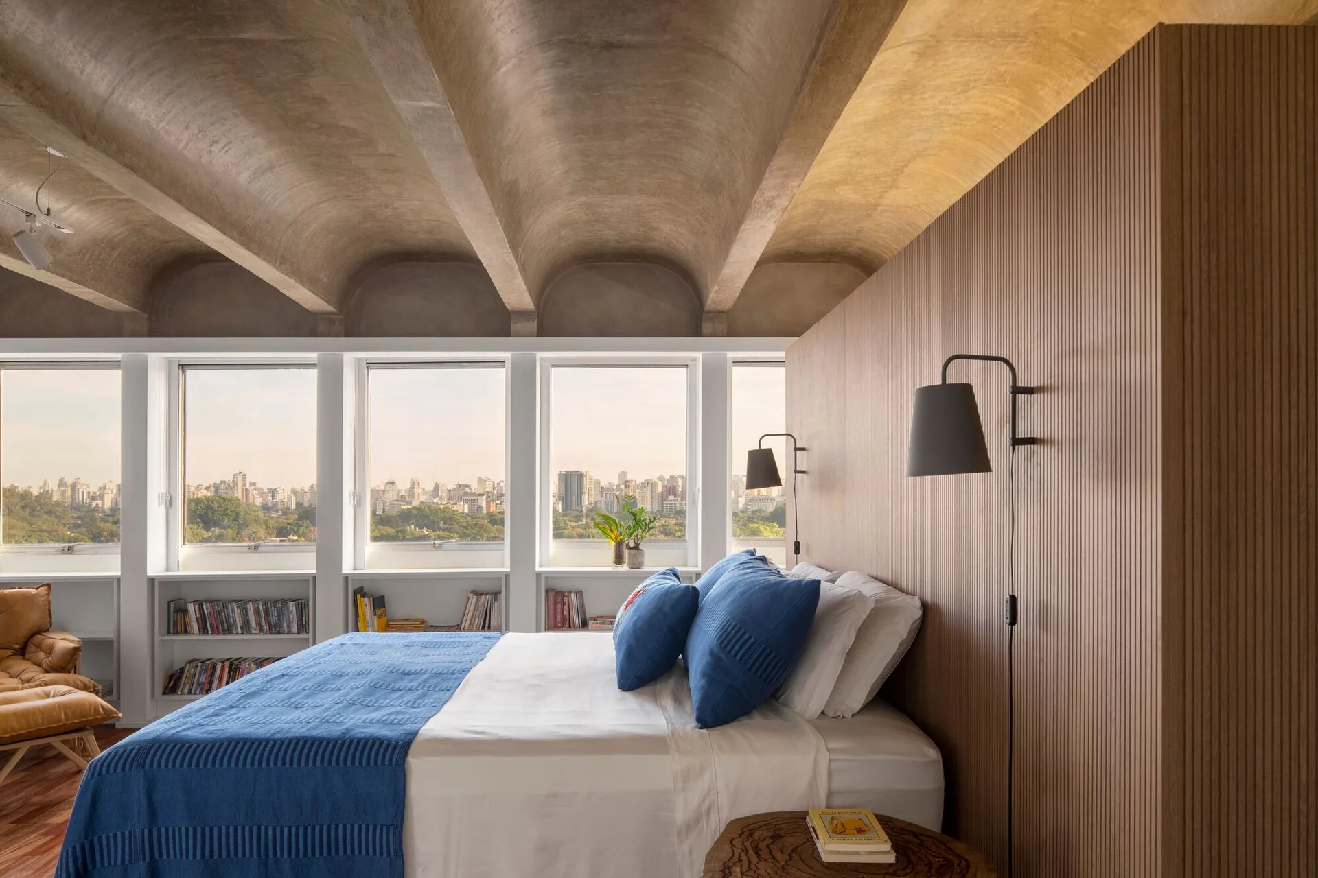 A modern bedroom with a concrete barrel-vaulted ceiling, a vertical wood slat accent wall, and built-in bookshelves beneath a row of windows.