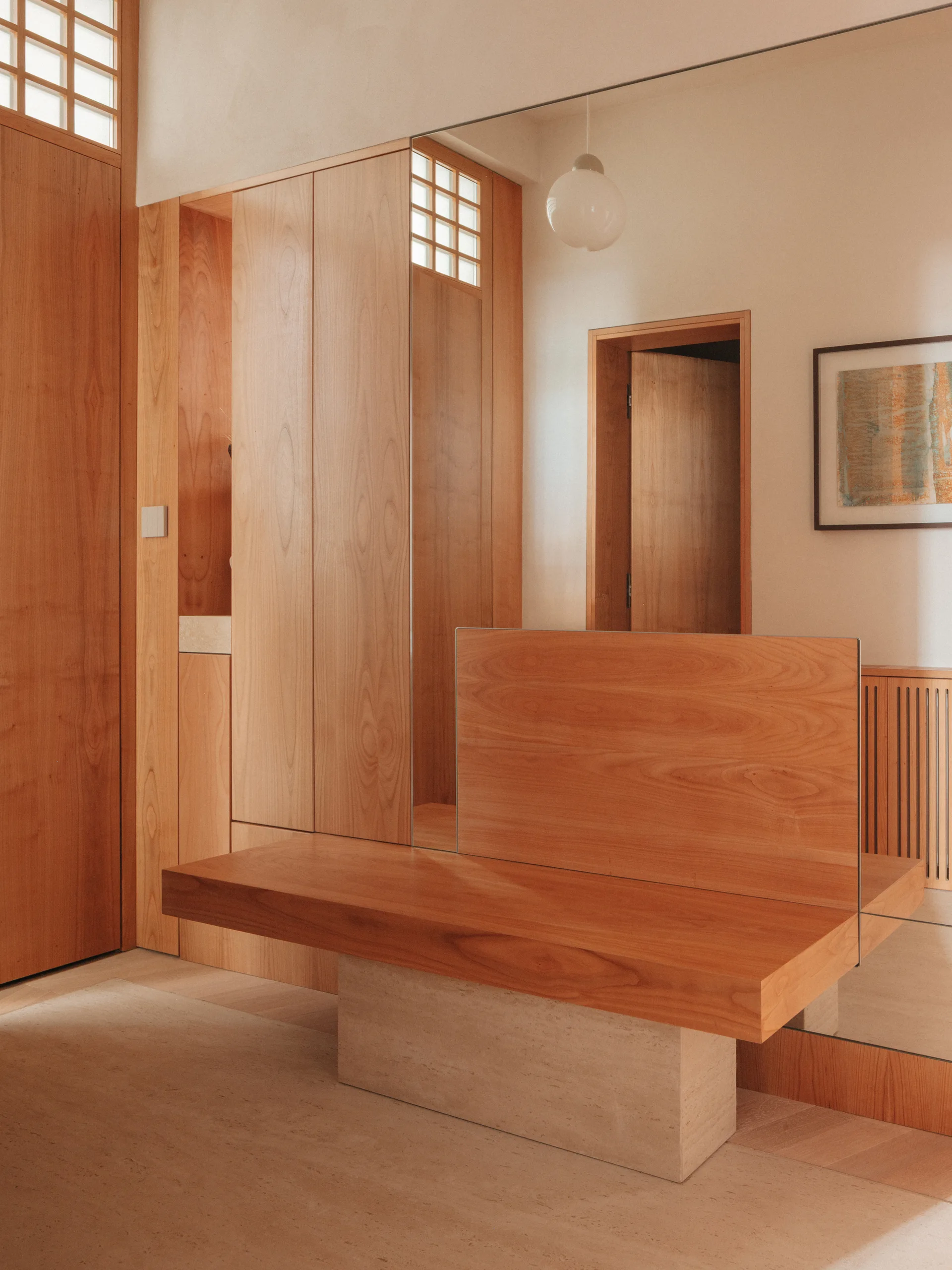A minimalist entryway features warm wood-paneled walls, integrated storage, and a wooden bench supported by a thick beige stone pedestal.
