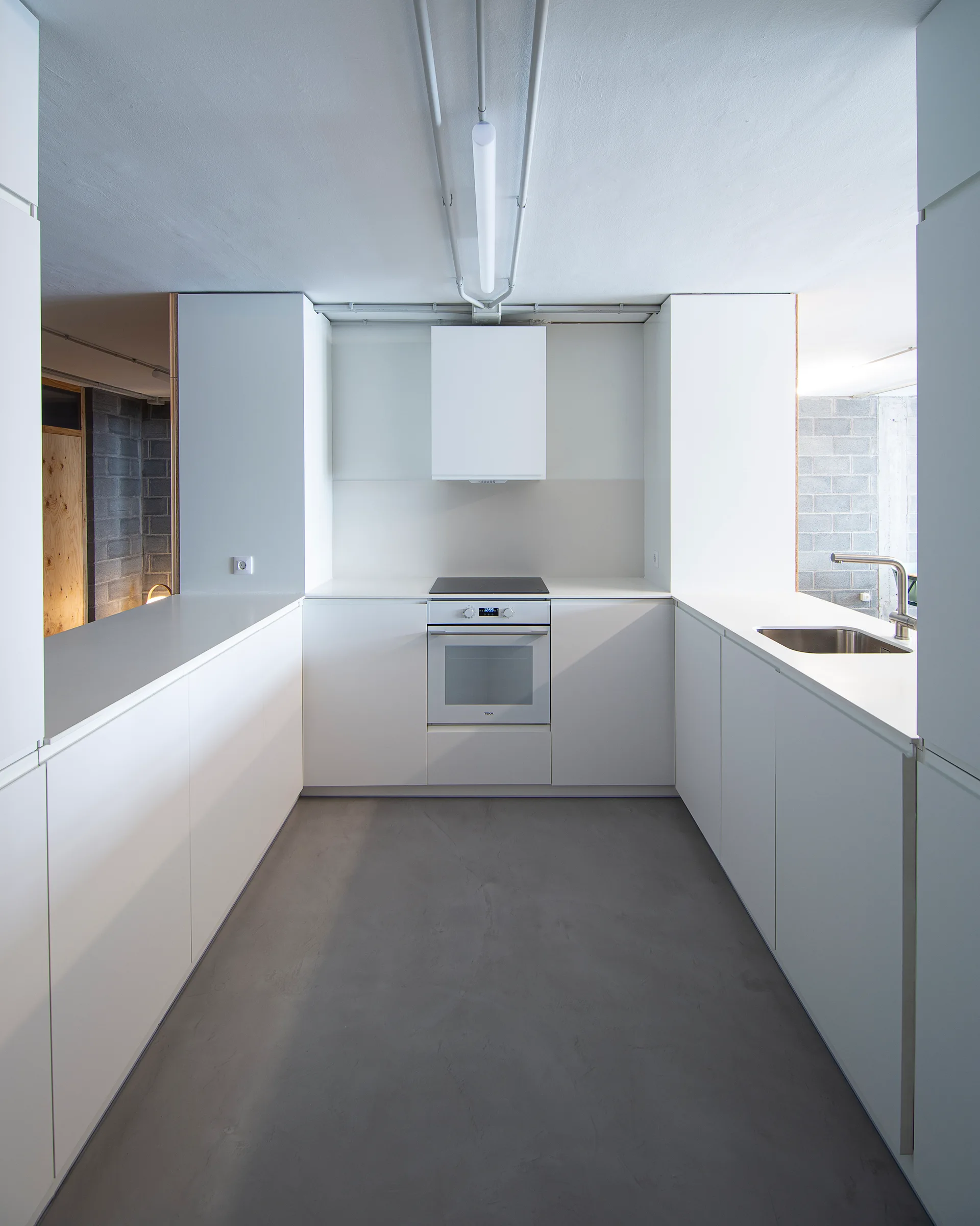 A minimalist U-shaped kitchen featuring matte white cabinetry, an integrated oven, stainless steel sink, and a smooth gray concrete floor.