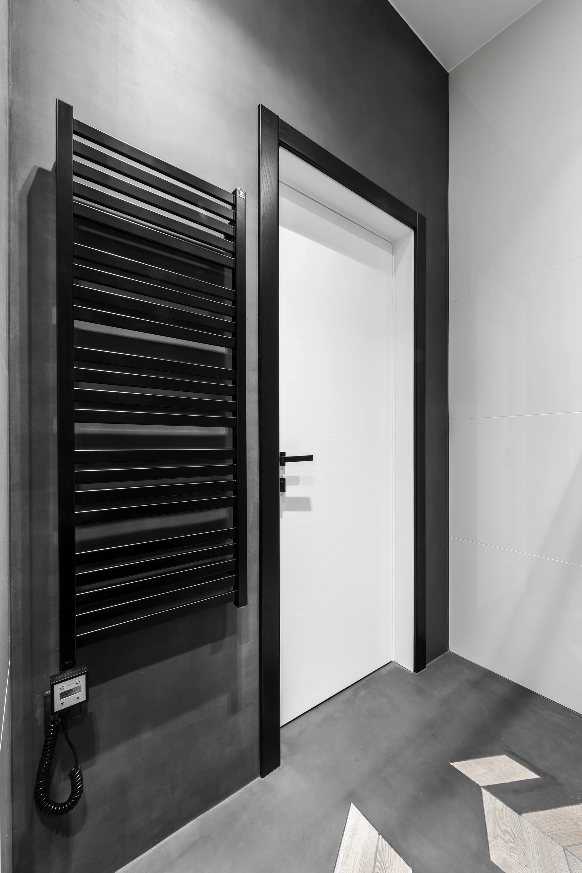A black wall-mounted ladder towel warmer hangs beside a white door with a bold black frame against a dark grey matte wall.