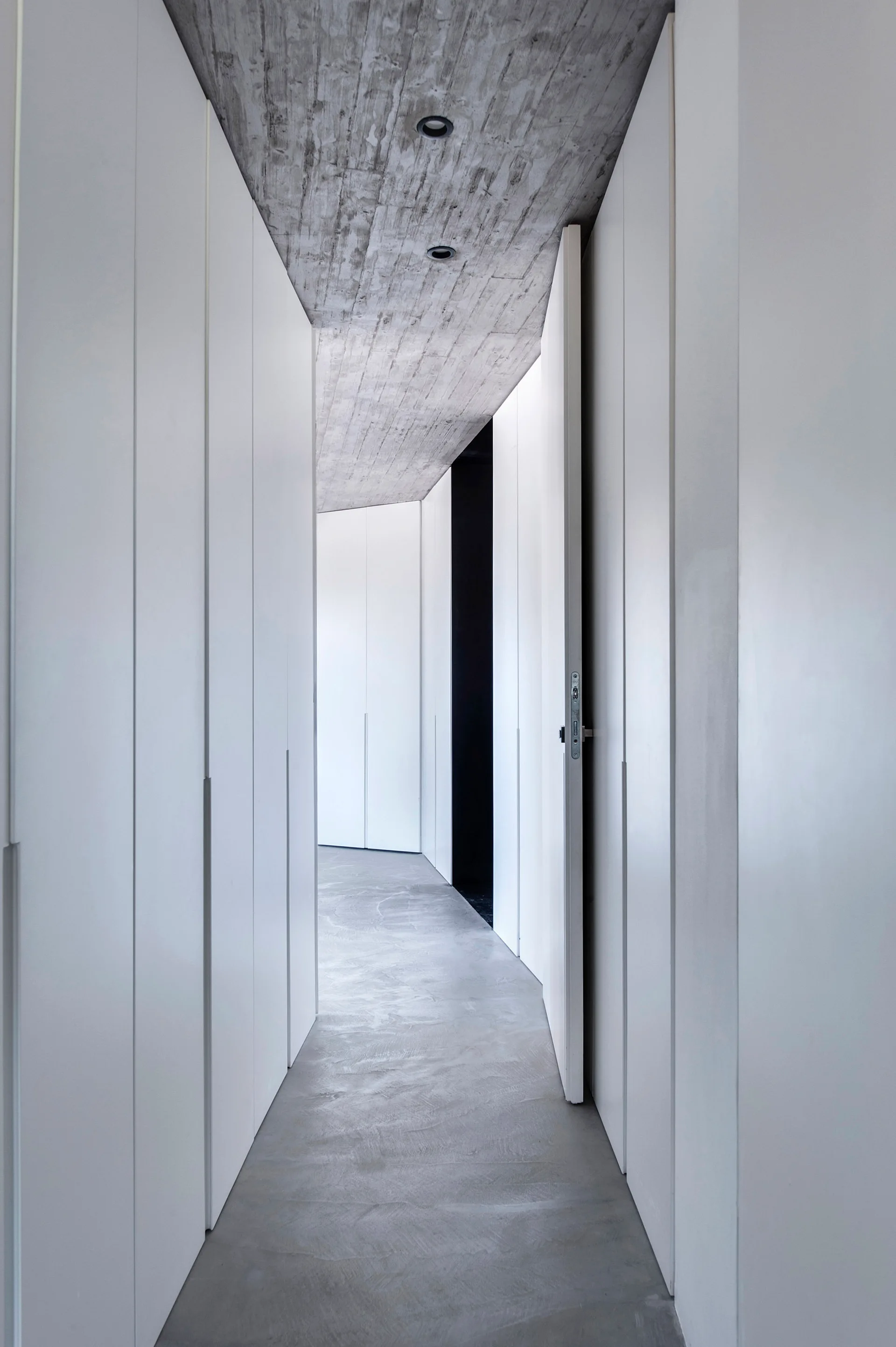 A narrow minimalist hallway features continuous flat white floor-to-ceiling cabinetry set beneath a raw textured concrete ceiling and floor.