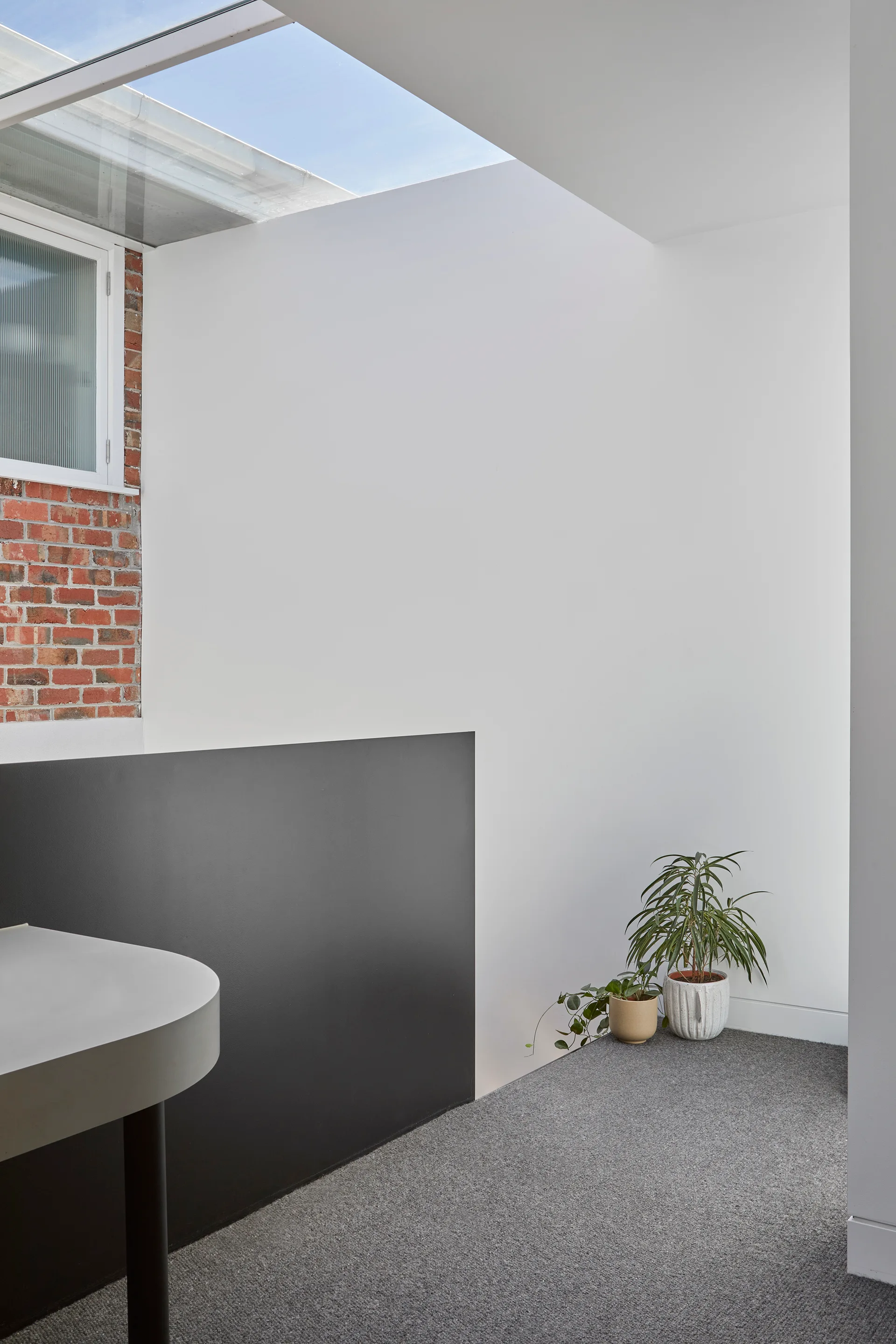 A minimalist hallway landing with gray carpet, a dark partition, a section of exposed red brick, and a glass skylight.