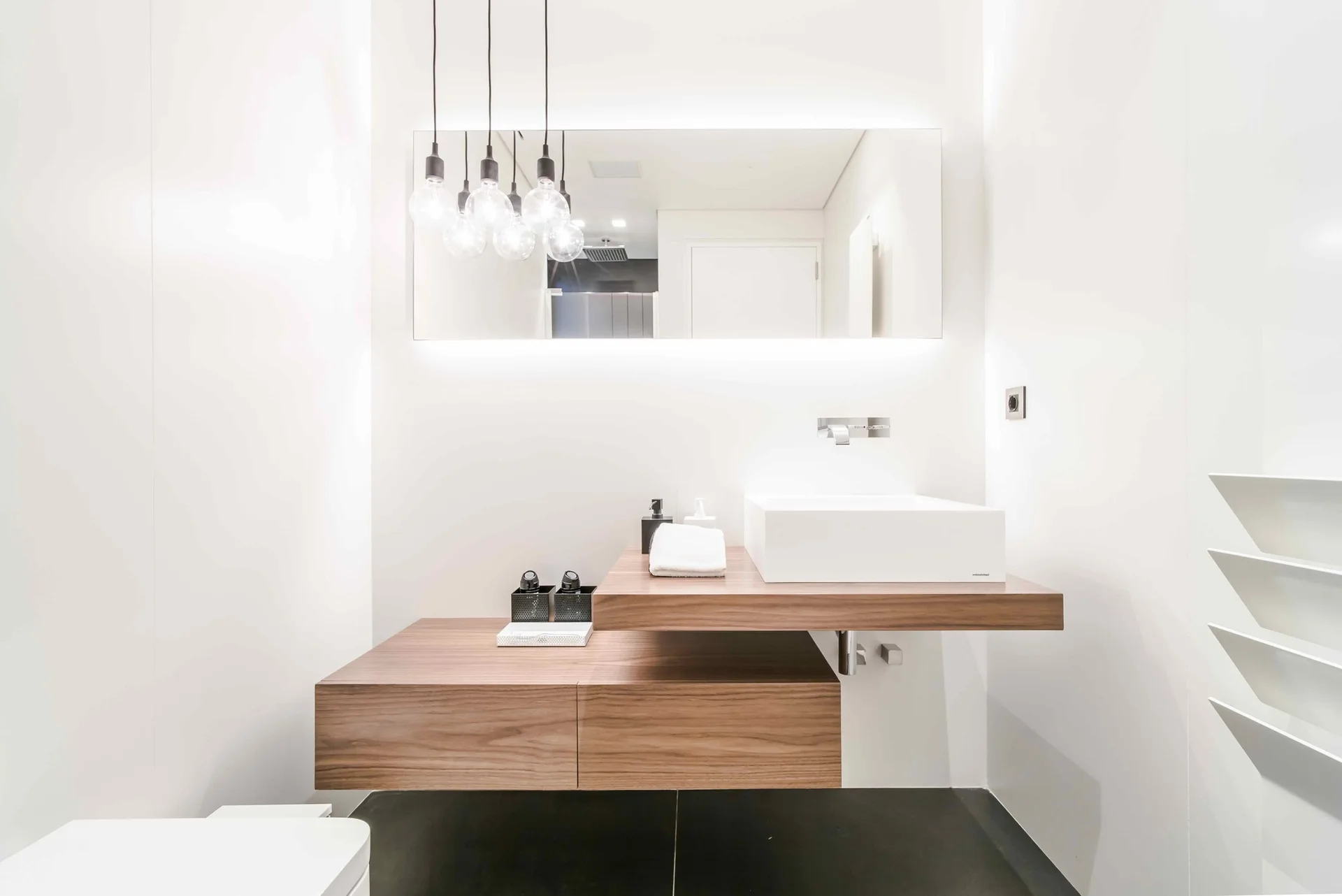 A minimalist powder room features a staggered floating wooden vanity, a white rectangular vessel sink, and a large backlit wall mirror.