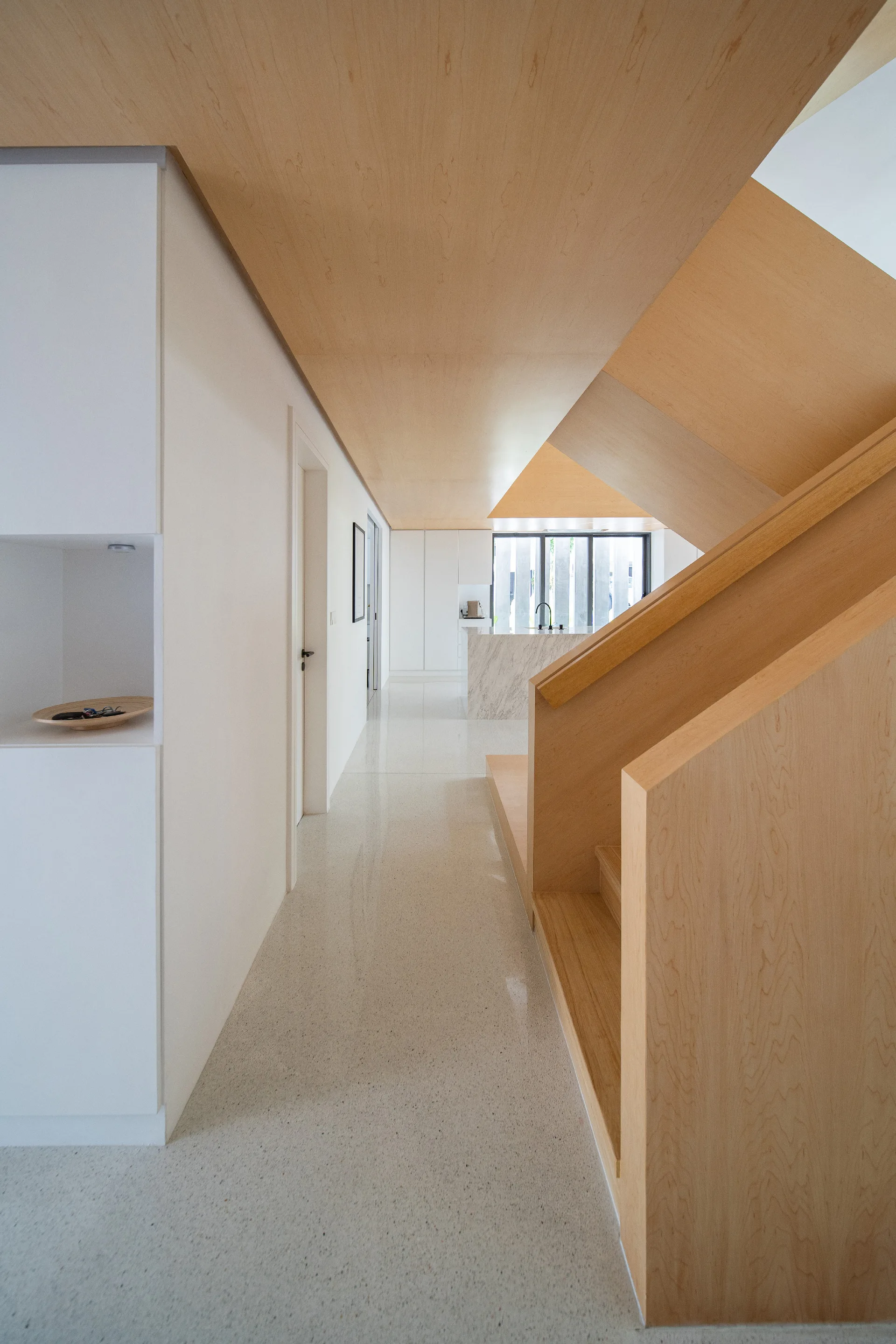 A modern minimalist hallway features a light wood paneled staircase, matching wood ceiling paneling, white walls, and speckled terrazzo flooring.