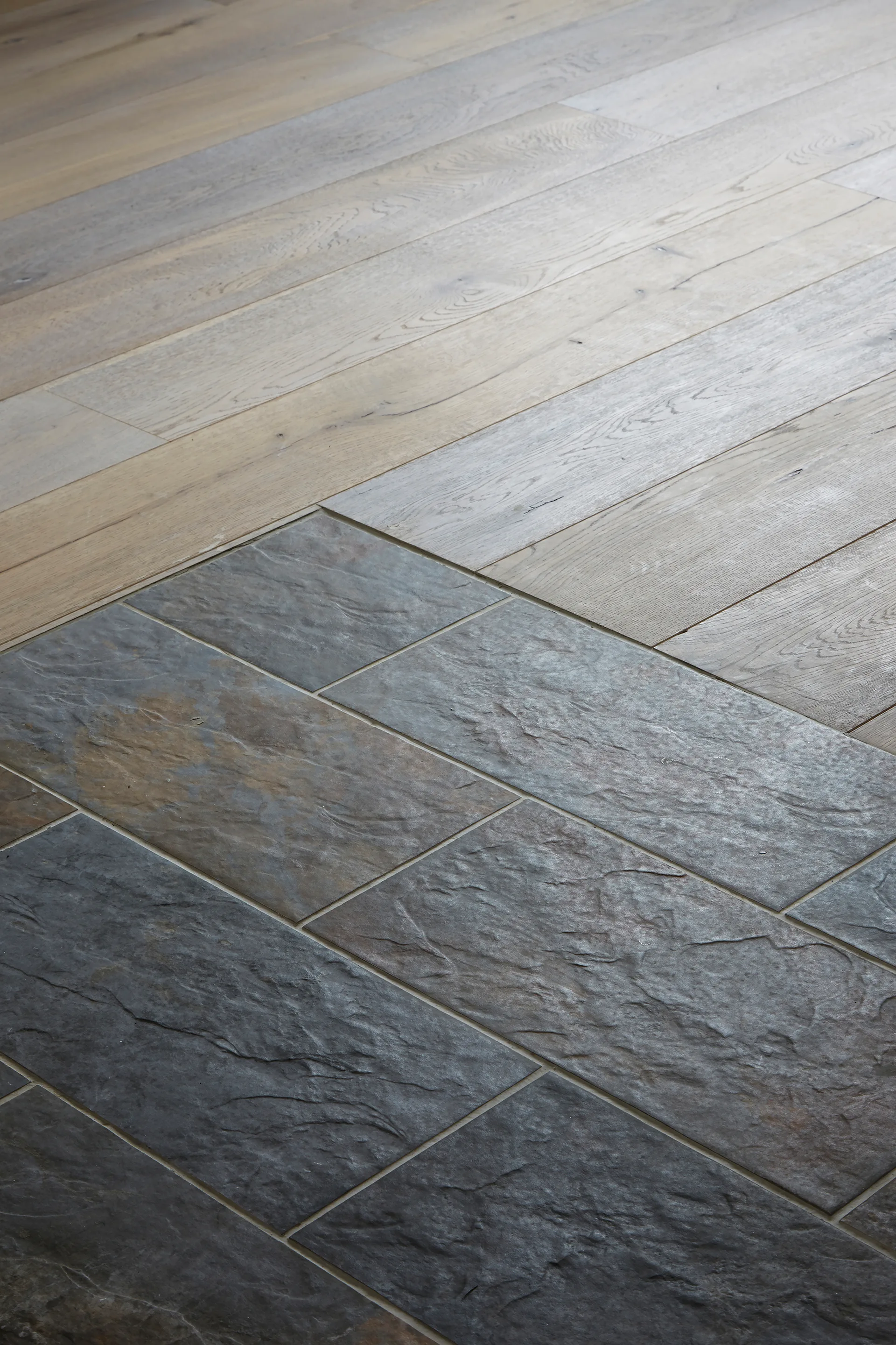 A close-up view of a floor transition where light oak wood planks meet textured dark grey slate tiles.