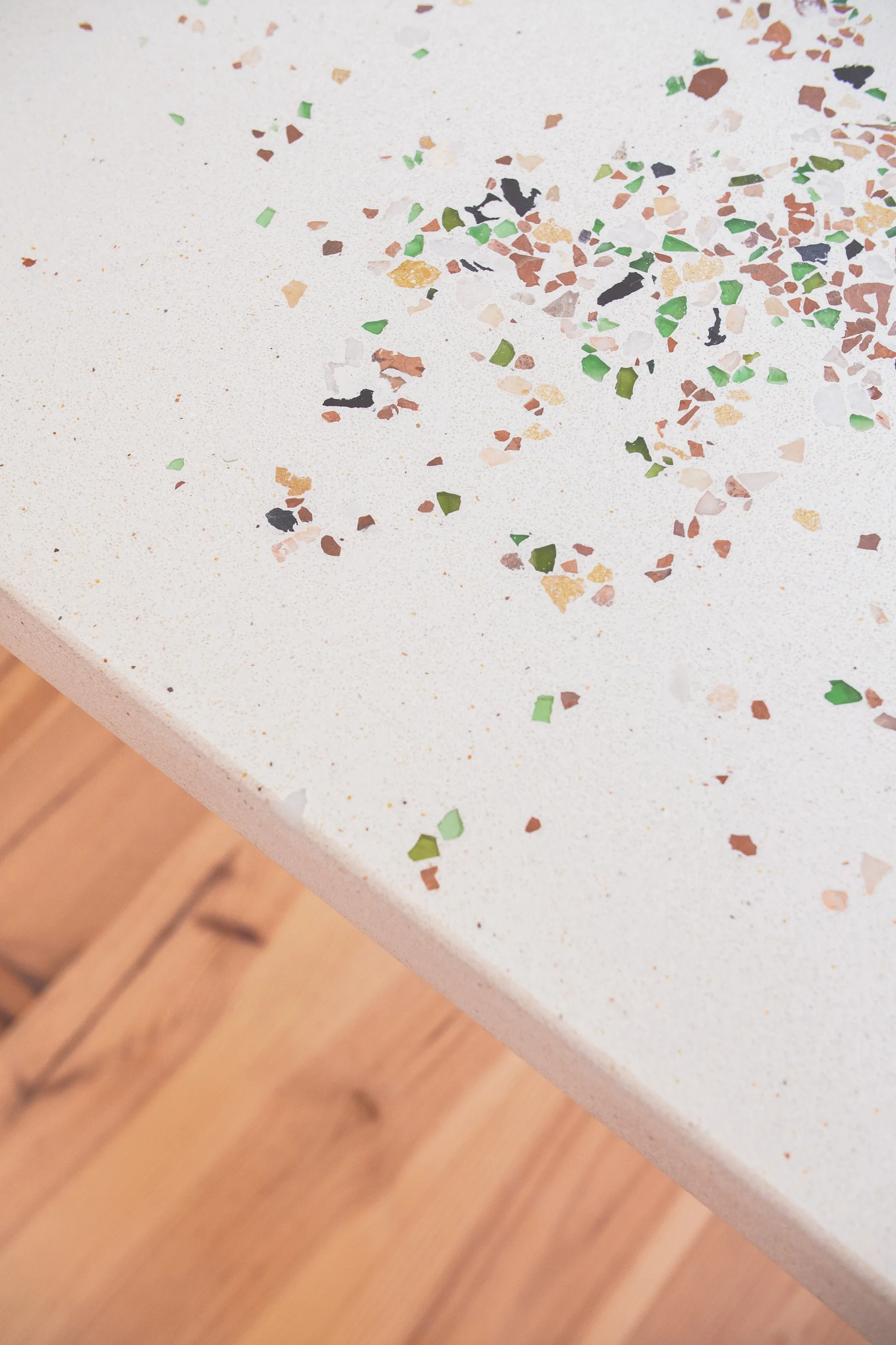 A close-up view of a white terrazzo countertop with embedded green and brown aggregate fragments above a light wood floor.