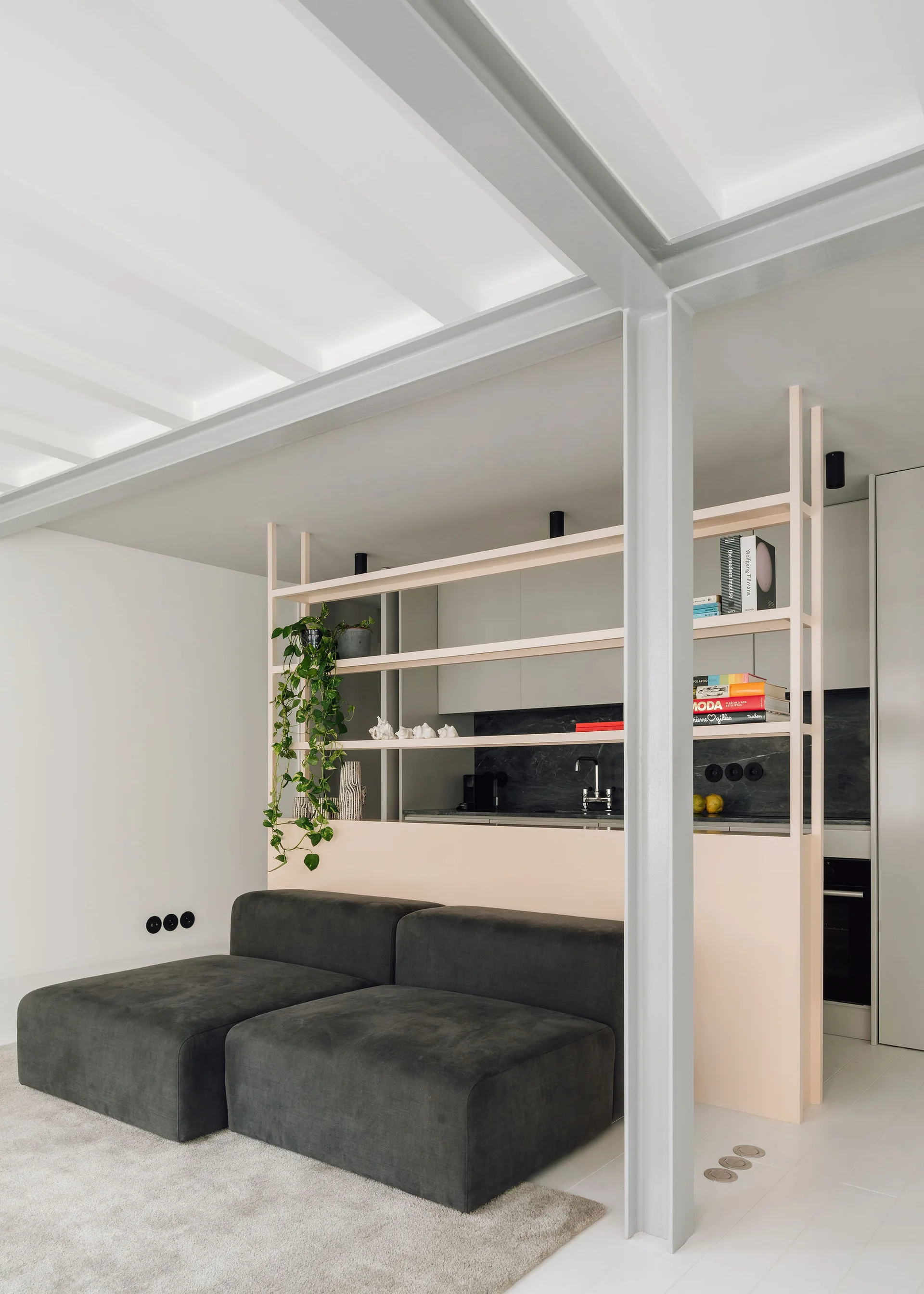 A minimalist living room features a dark grey sectional sofa and a soft pink open-shelf divider.