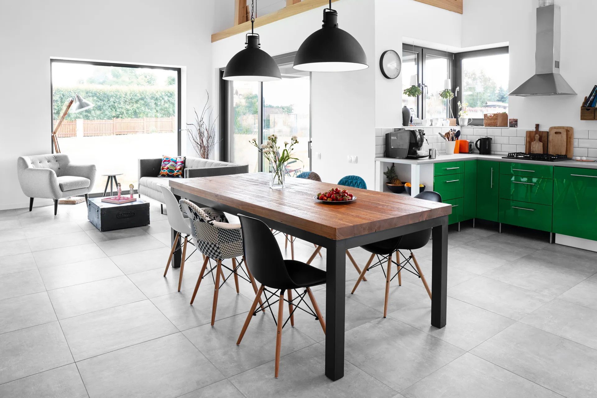 An open-concept dining space features a rustic wood table with mismatched chairs, large black dome pendant lights, and glossy green kitchen cabinets.