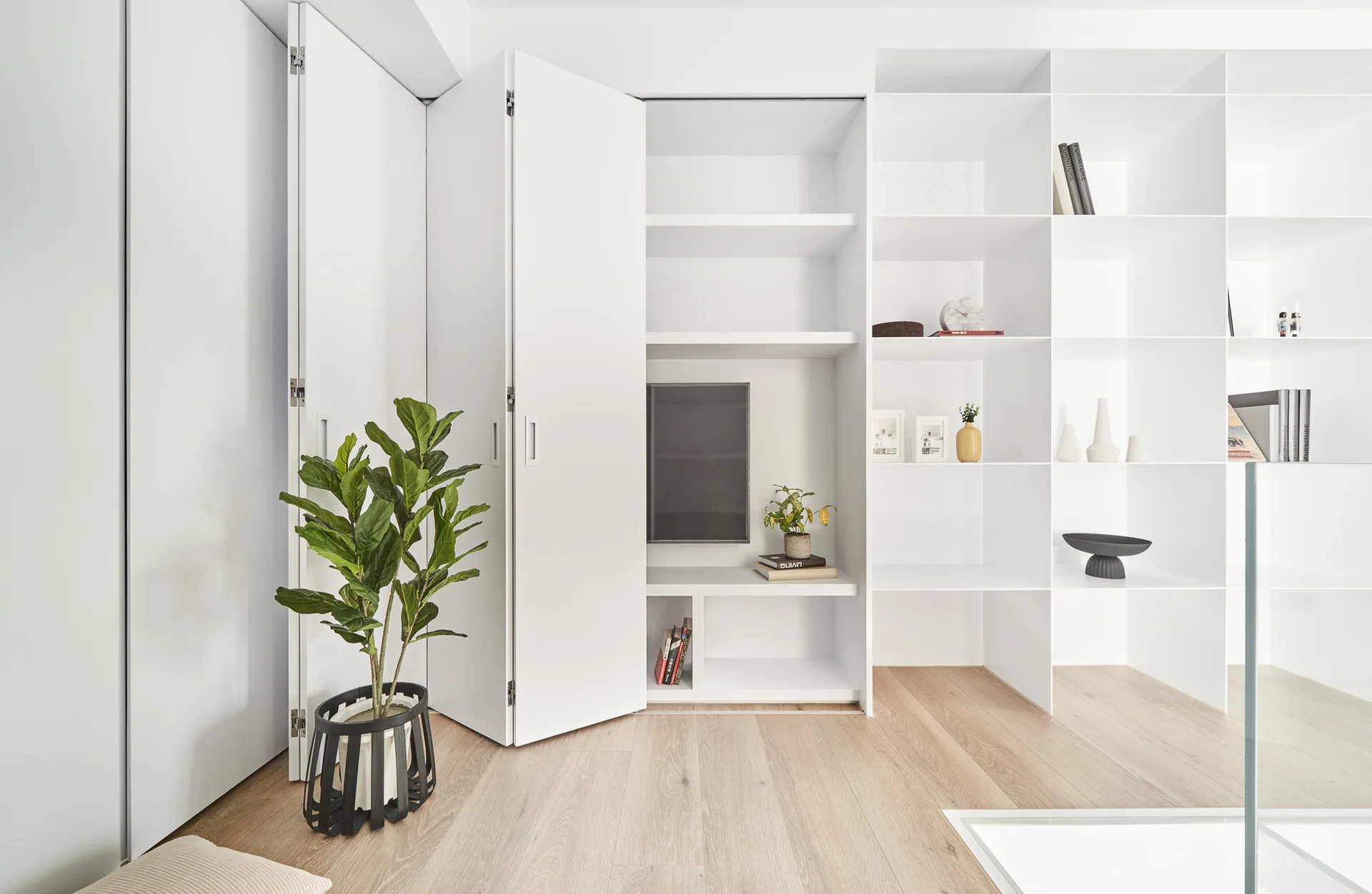 White built-in shelving and cabinetry with partially opened bi-fold doors on a light wood floor next to a green potted plant.