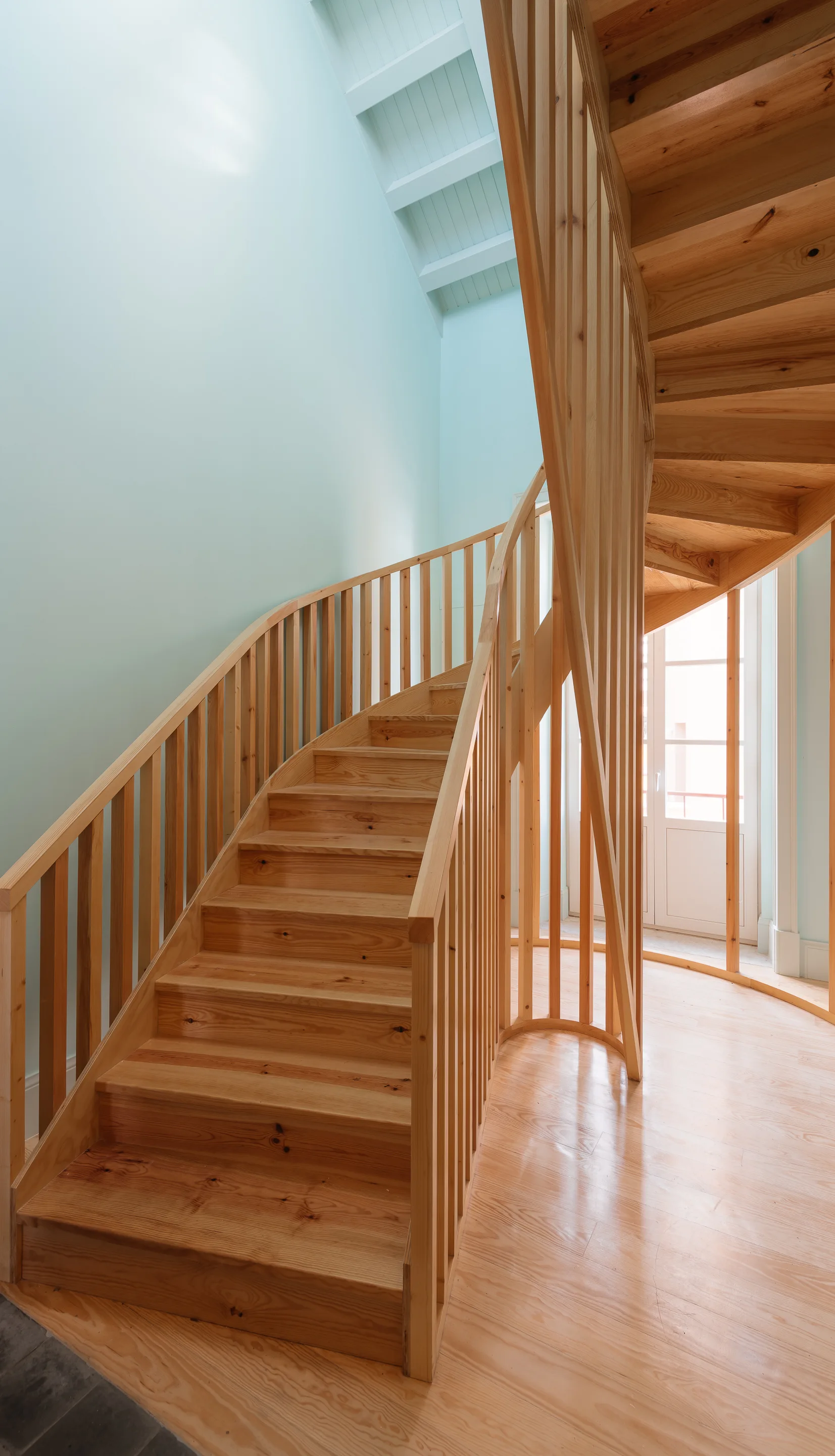A light wood spiral staircase with a vertical slat railing stands against pale mint green walls beneath a bright skylight.
