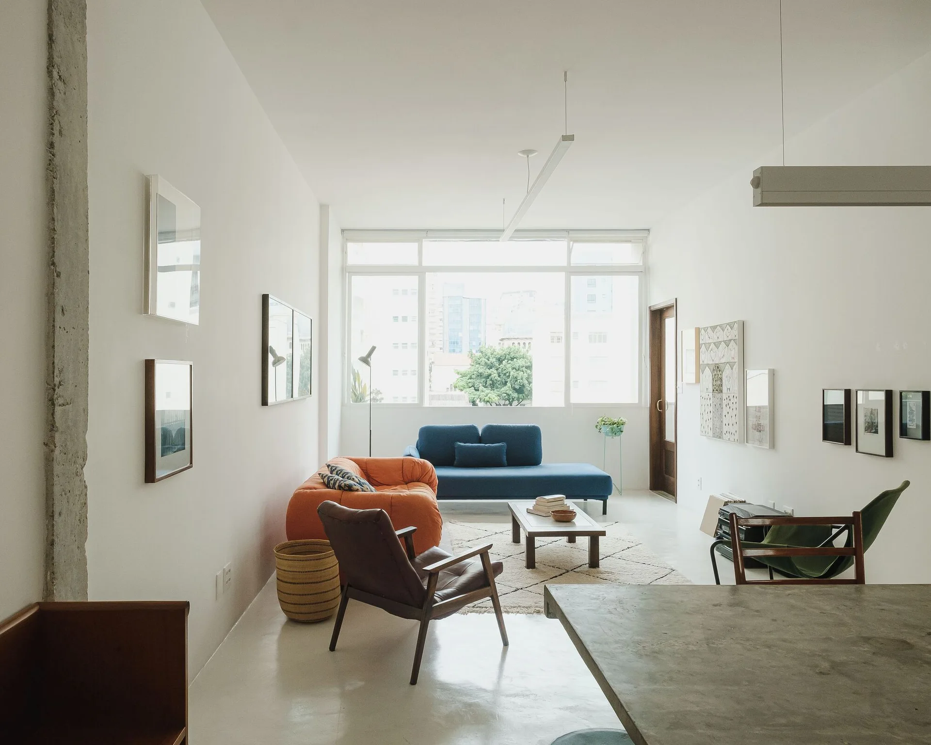 A bright minimalist living room features an orange sofa, a blue chaise, a leather armchair, and a large window overlooking city buildings.