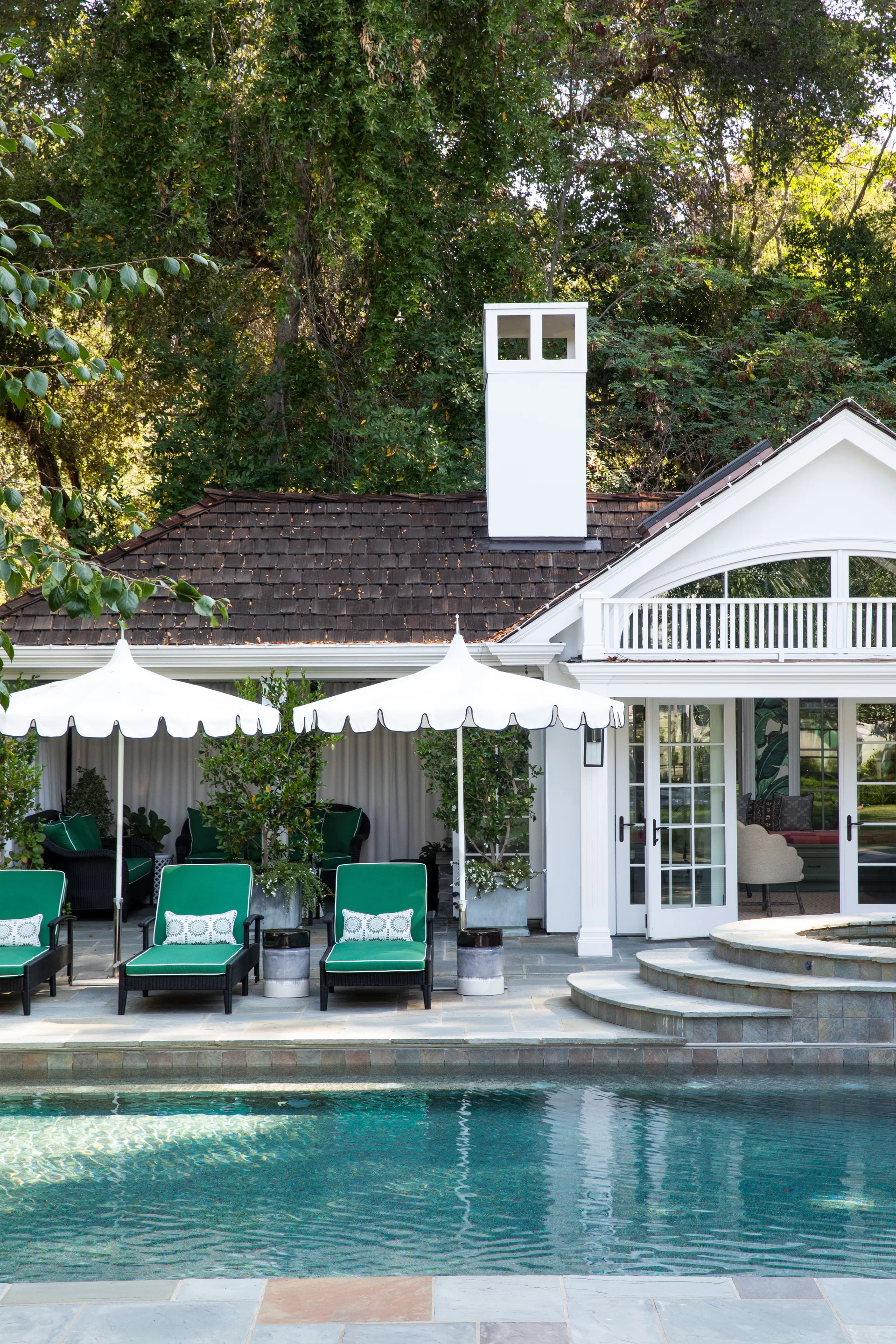 A backyard swimming pool features emerald green chaise lounges with scalloped white umbrellas on a stone patio beside a white cabana.