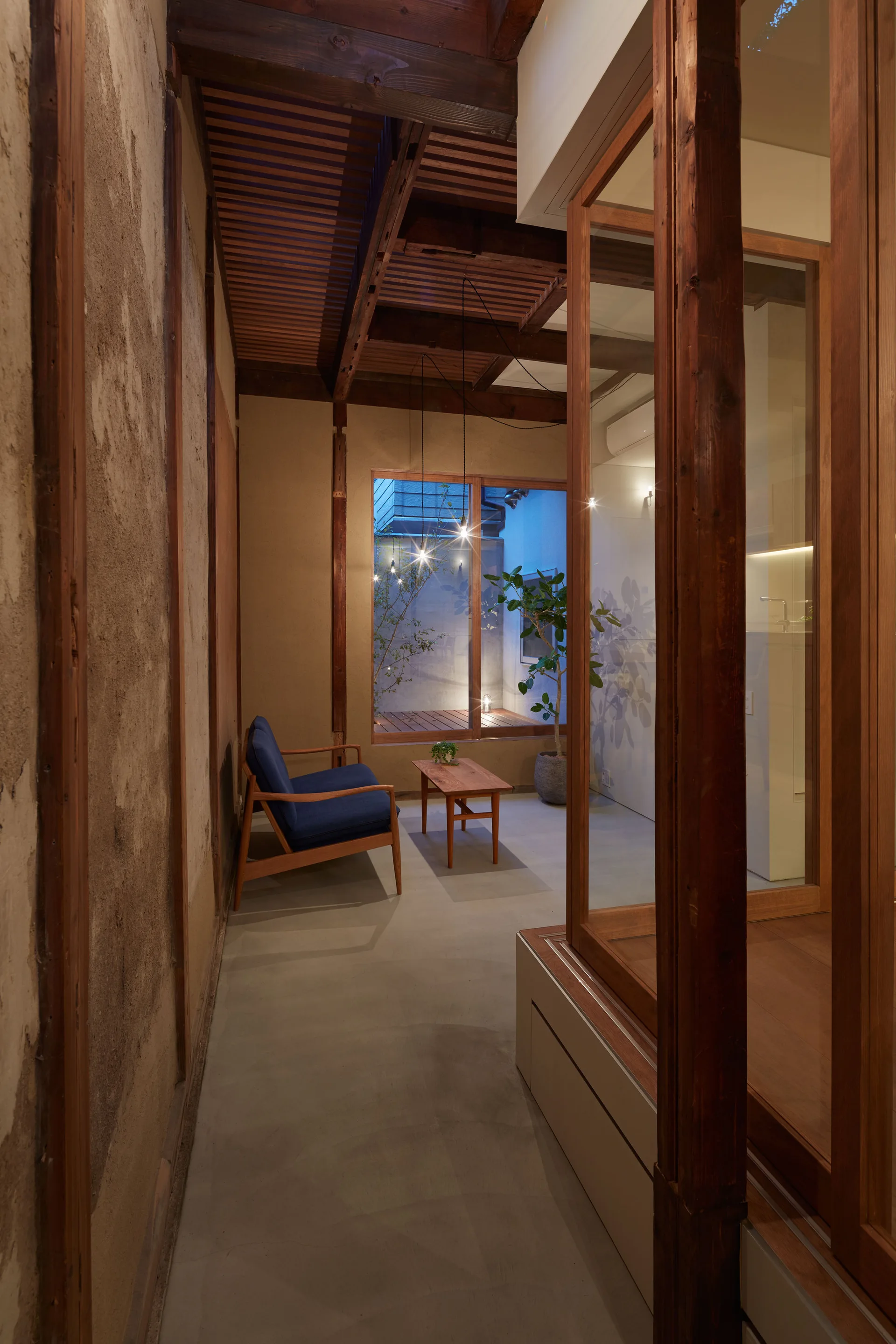 A minimalist living space with a blue wooden armchair, a low coffee table, textured plaster walls, and a large window looking out to a courtyard.