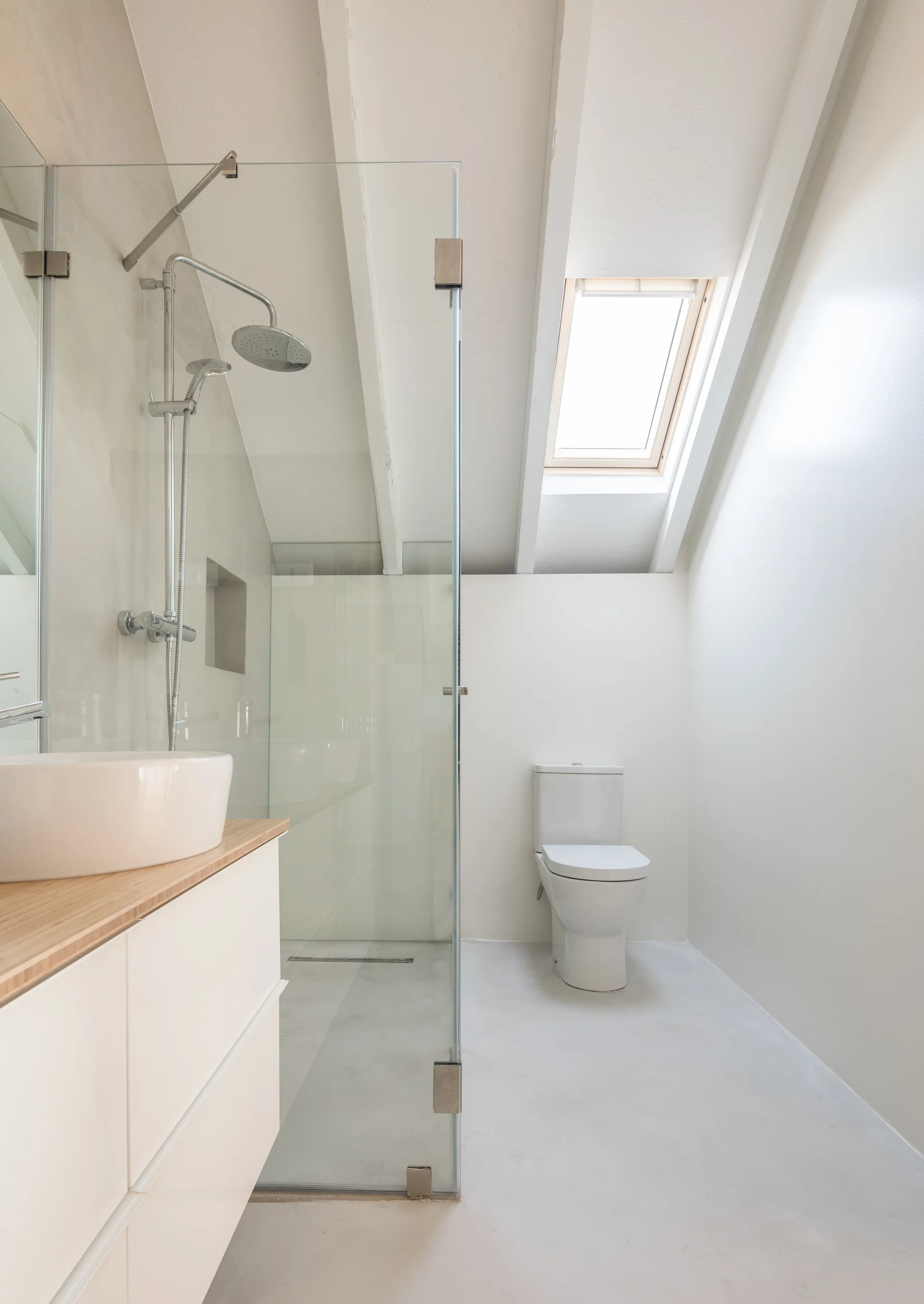 A minimalist attic bathroom with a white vanity, a clear glass shower, and a toilet positioned under a slanted ceiling with a skylight.