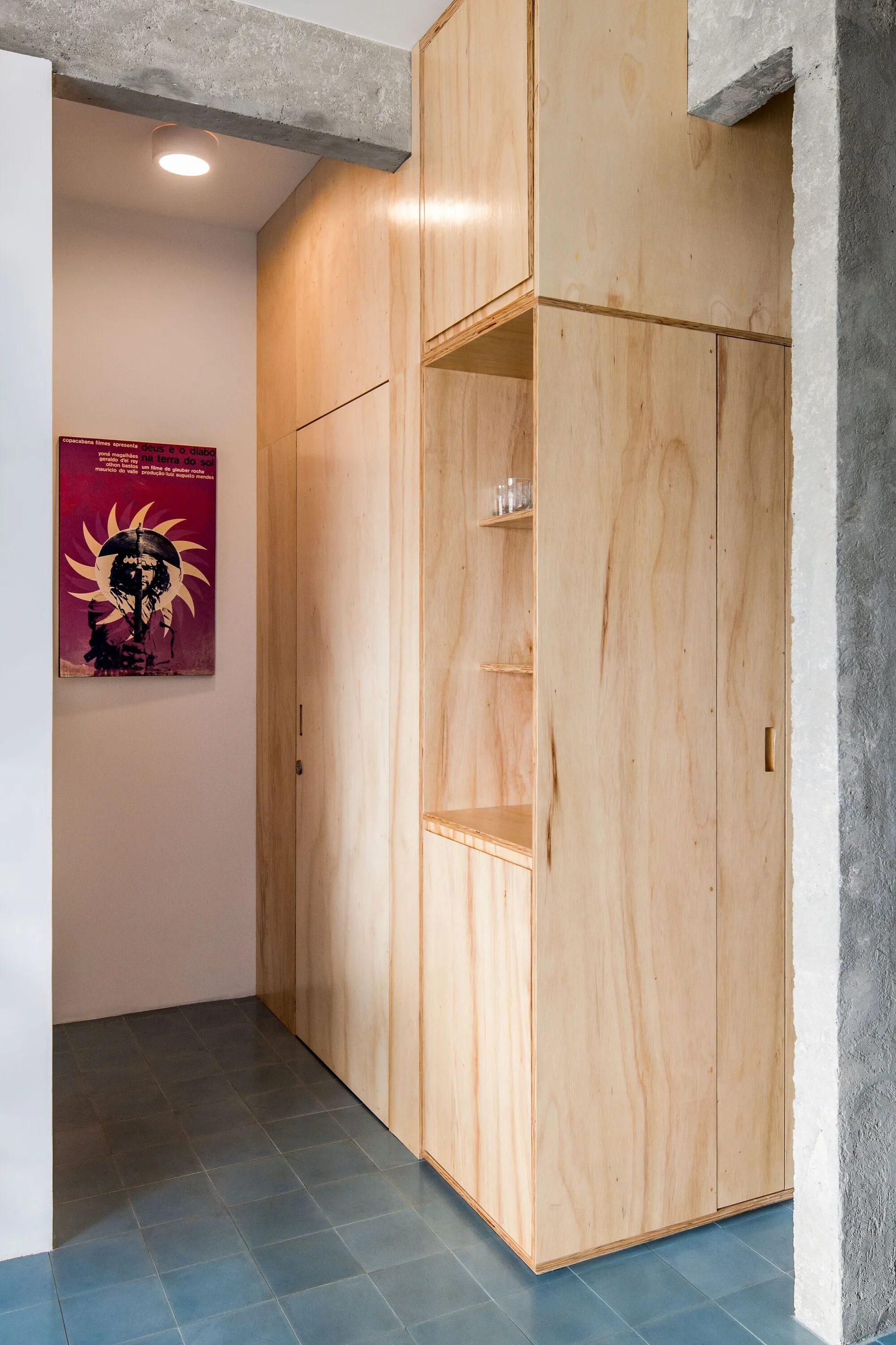 A hallway features a floor-to-ceiling plywood storage unit, blue tile flooring, an exposed concrete ceiling beam, and a purple poster on a white wall.