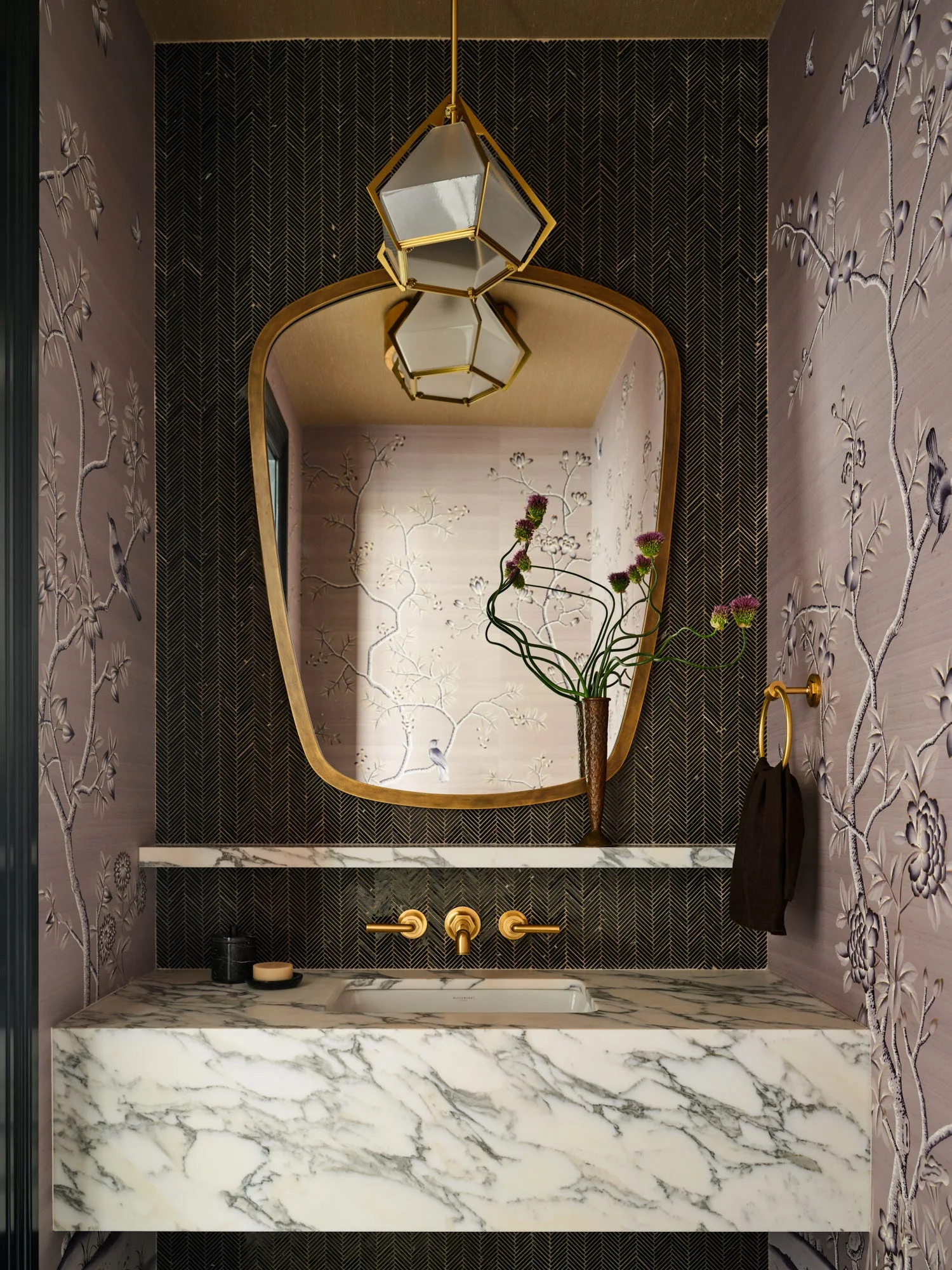 A powder room vanity features a veined marble countertop, wall-mounted gold faucet, black herringbone tile backsplash, and floral wallpaper.