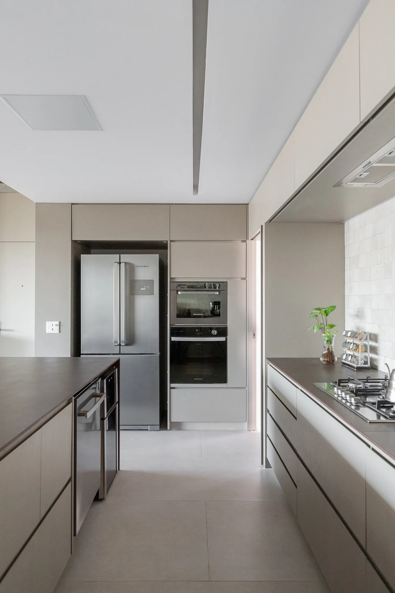 A modern galley kitchen features matte taupe handleless cabinets, built-in stainless steel appliances, a dark countertop, and large gray floor tiles.