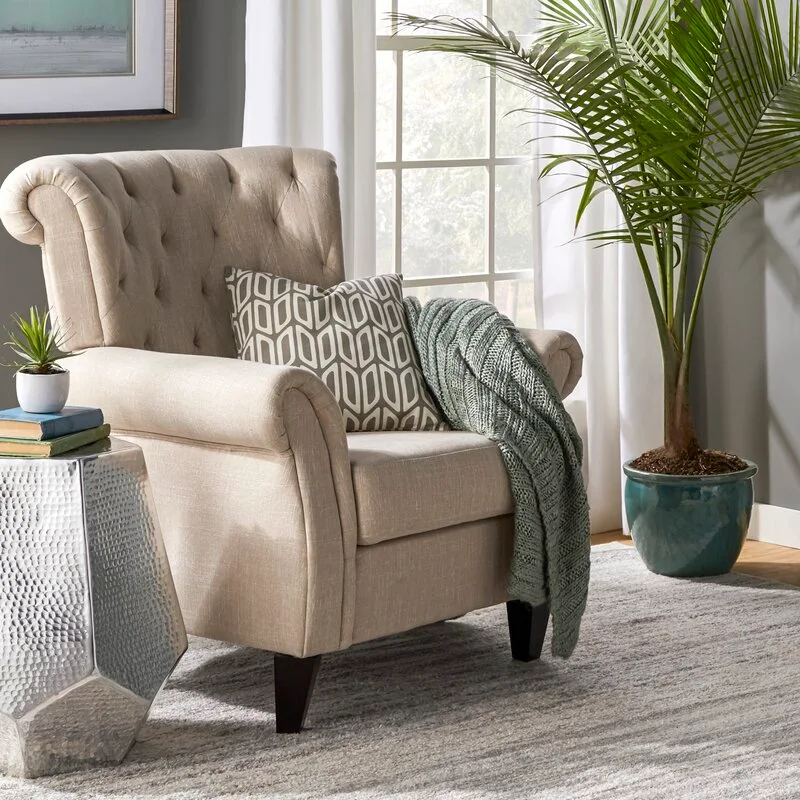A tufted beige wingback armchair sits next to a faceted silver side table, a tall potted palm, and a large window with white curtains.