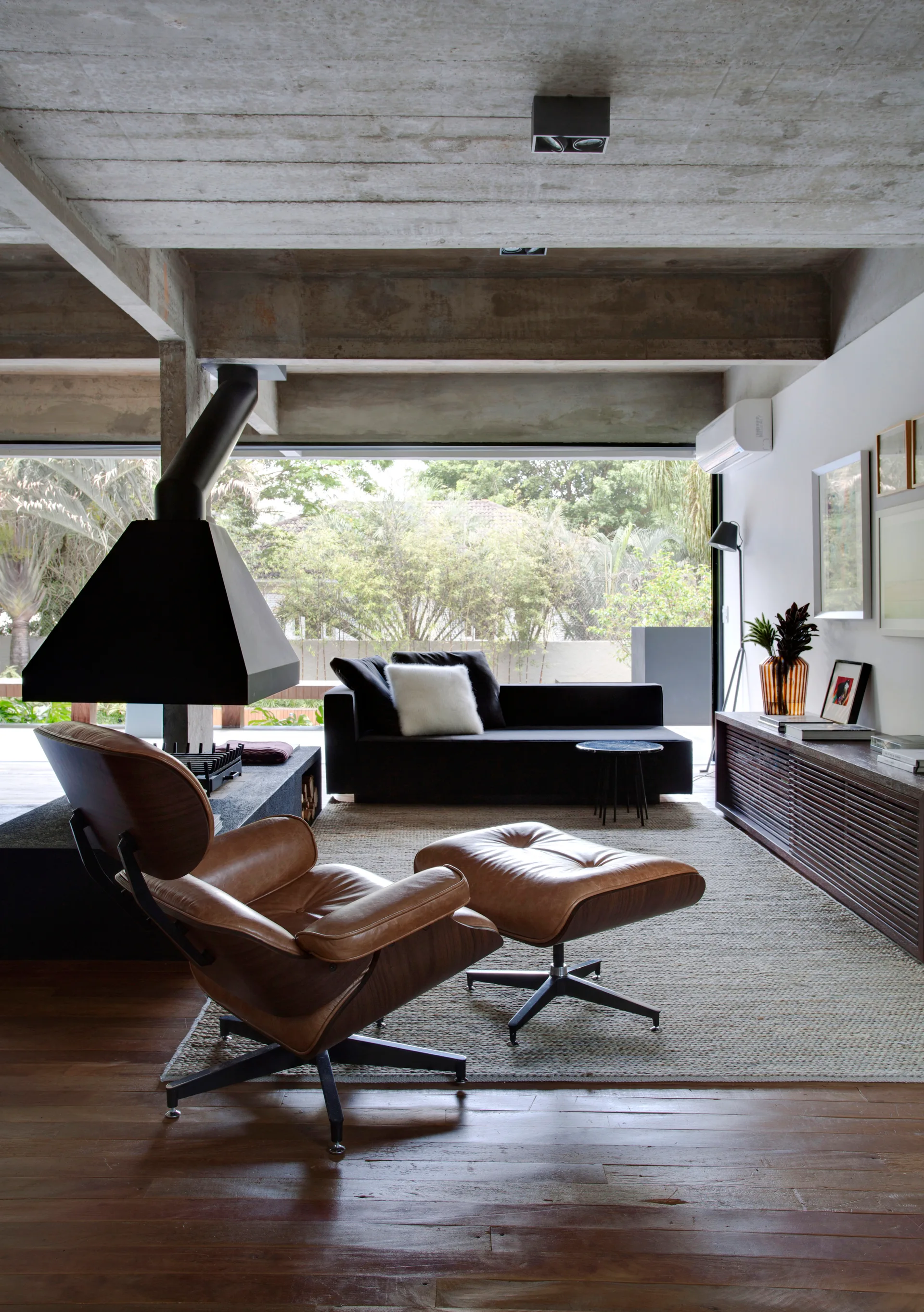 A living room featuring an exposed concrete ceiling, a leather lounge chair with ottoman, a suspended black fireplace, and a large sliding glass door.