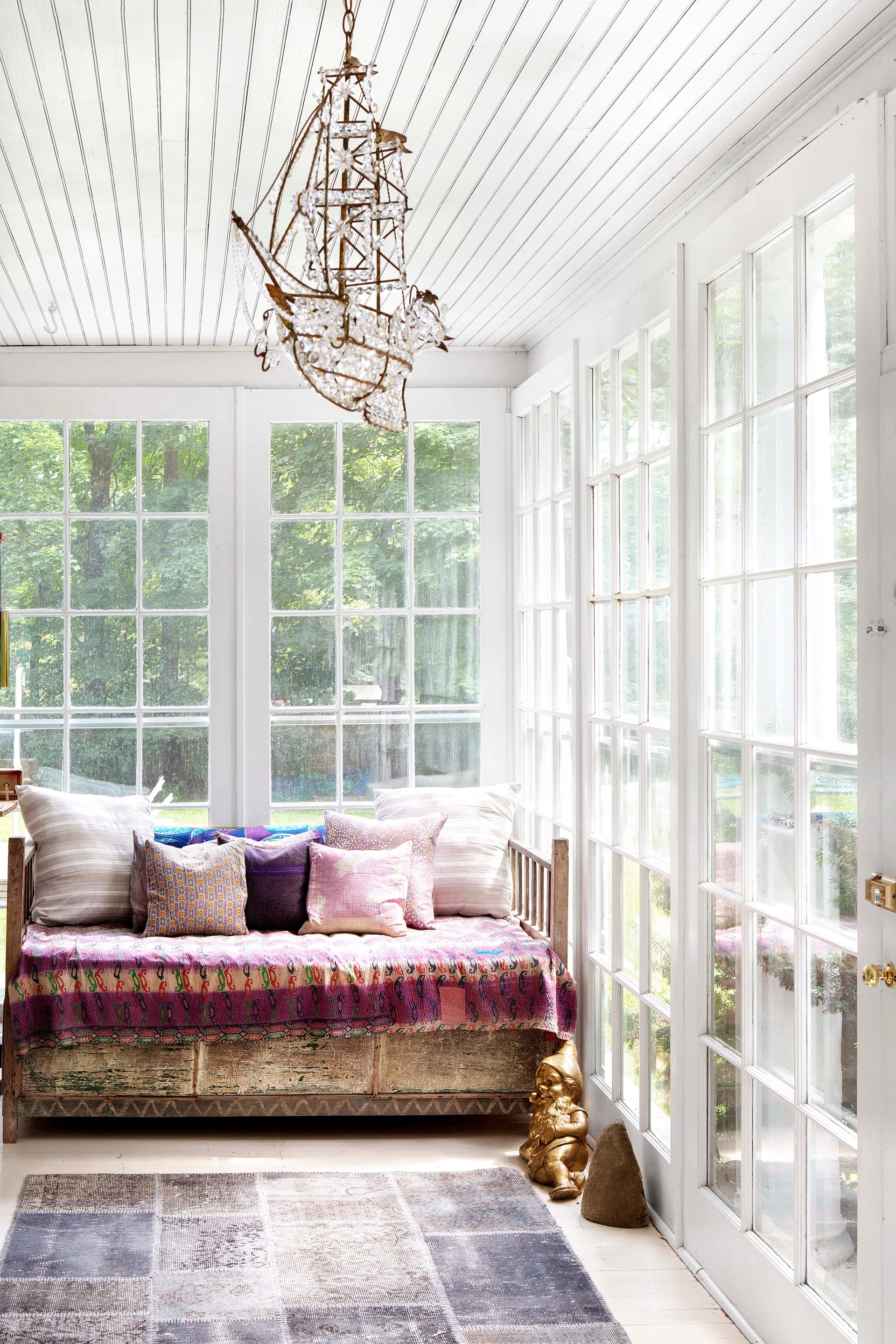 A sunroom with white paneled walls and ceiling features a wooden daybed with colorful pillows, a patterned rug, and a crystal ship chandelier.