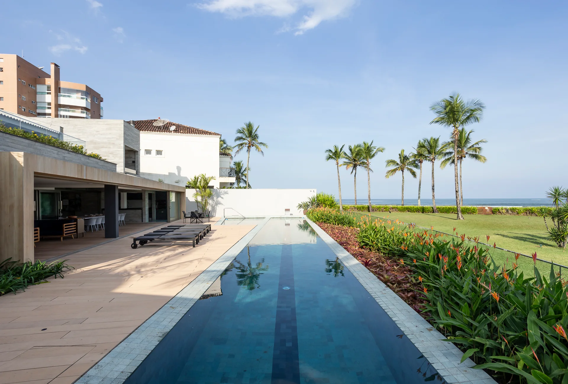 A long rectangular lap pool runs parallel to a wood-planked patio deck with lounge chairs next to a green lawn with palm trees.