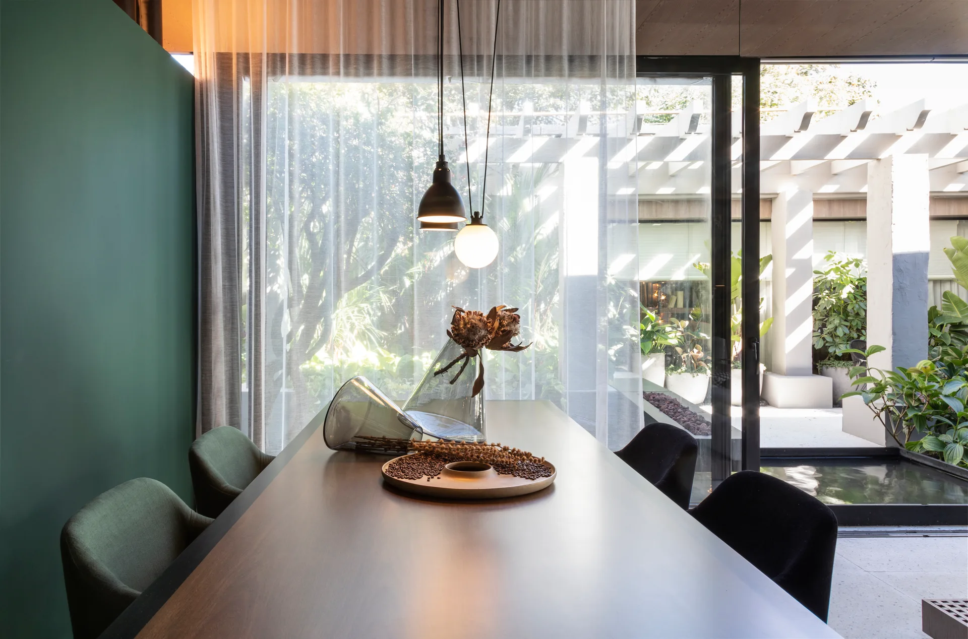 A modern dining room with a dark wood table and chairs situated next to sheer white curtains and a sliding glass door to a garden.
