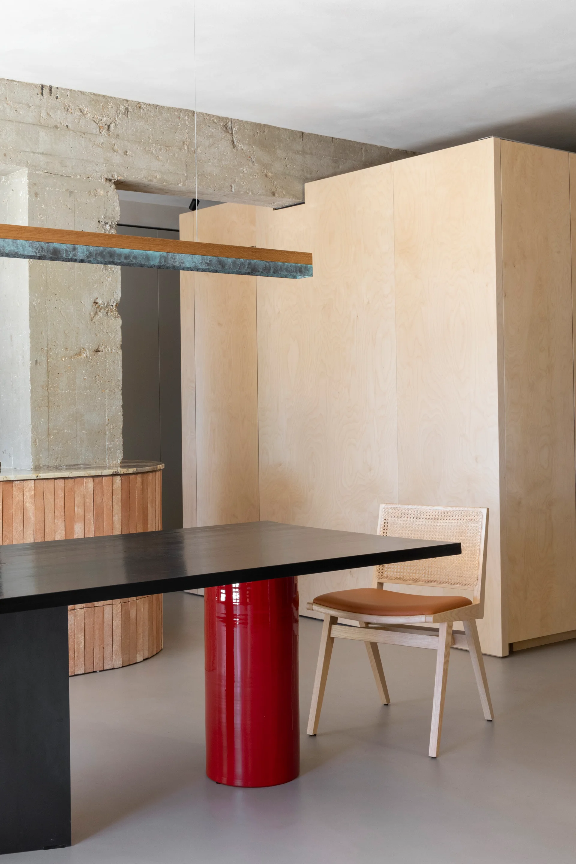 A modern black dining table with a glossy red pedestal base and a cane back chair sit beside a raw concrete column and light wood cabinetry.